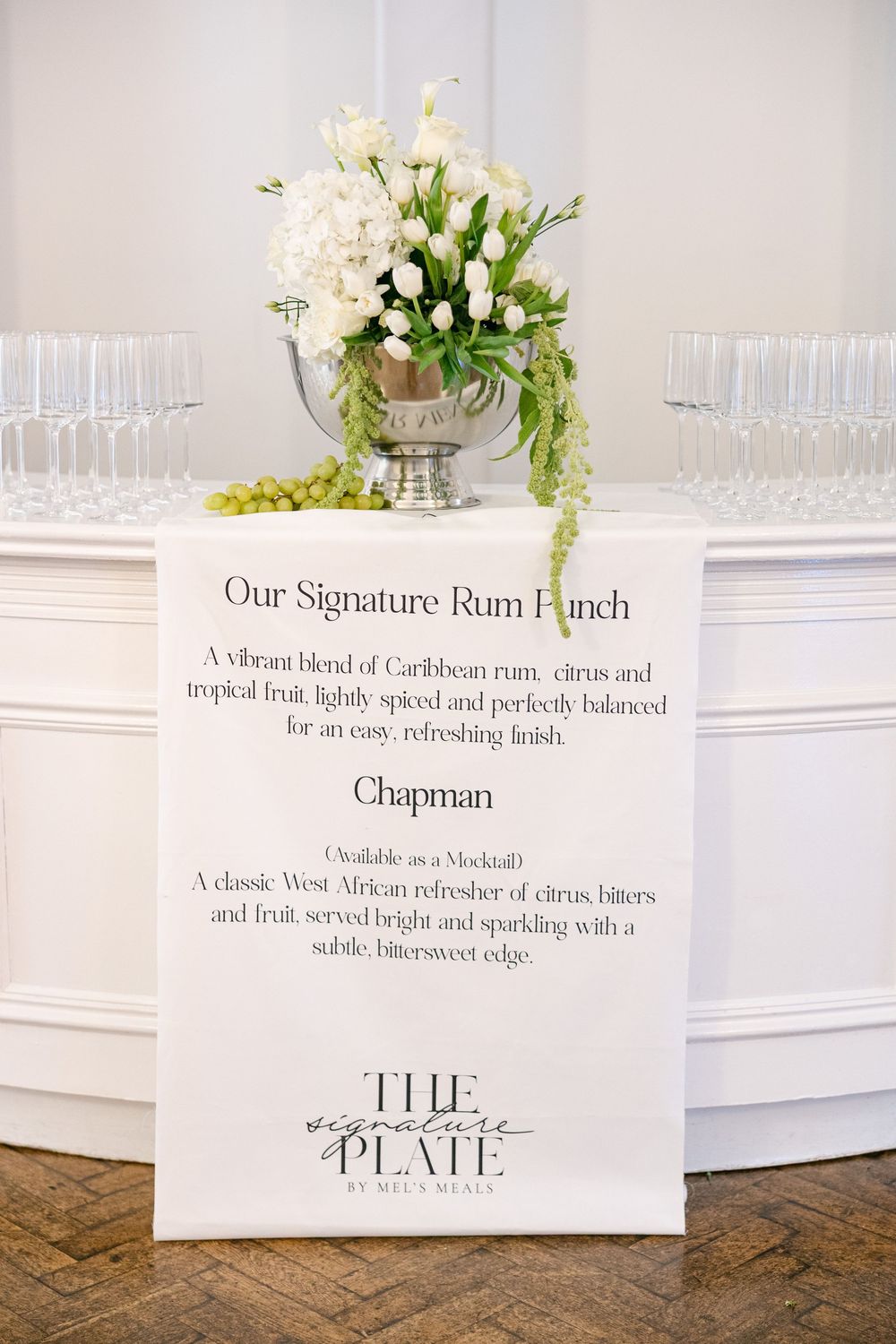 Elegant wedding bar menu sign for Signature Rum Punch and Chapman mocktail displayed beside white floral arrangement.