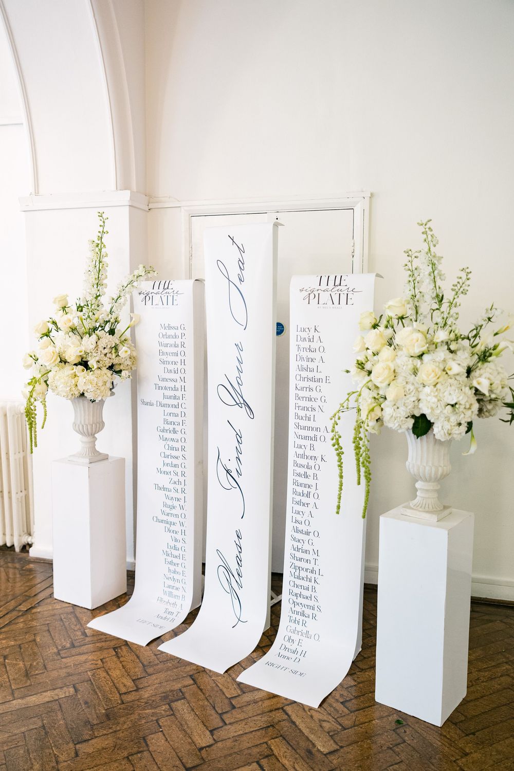 Elegant wedding seating chart displayed on tall white boards with calligraphy script, flanked by white floral arrangements in urns.