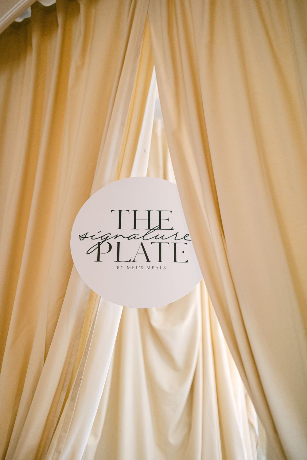The Signature Plate by Mels Meals logo sign displayed on cream draped fabric backdrop.