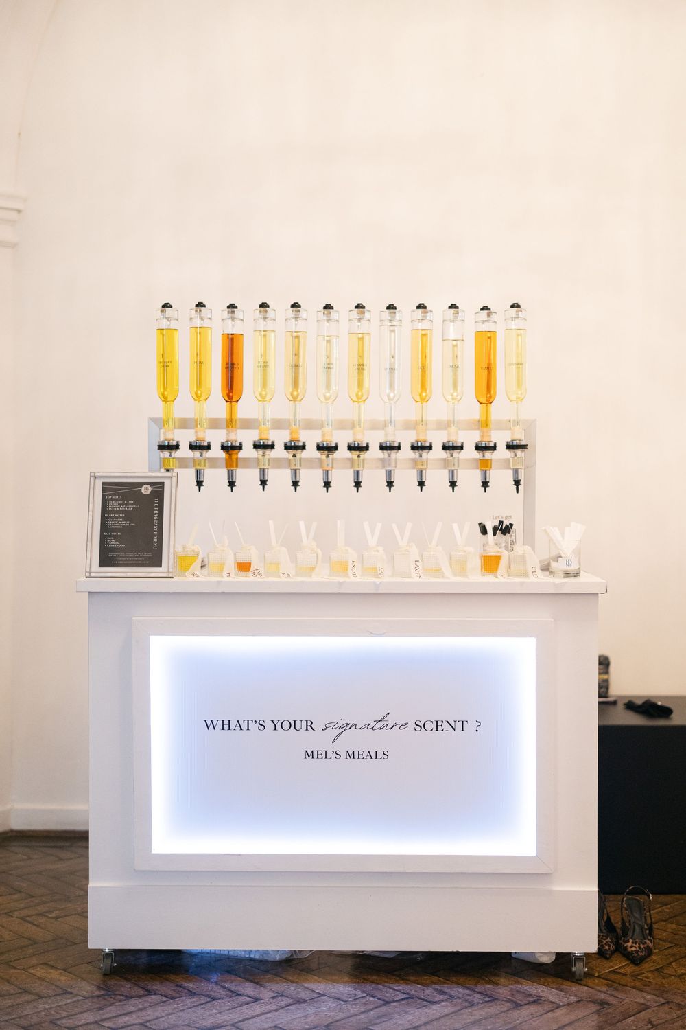 White illuminated perfume bar with syringes of fragrant oils displayed, reading What's Your Signature Scent? by Mel's Meals.