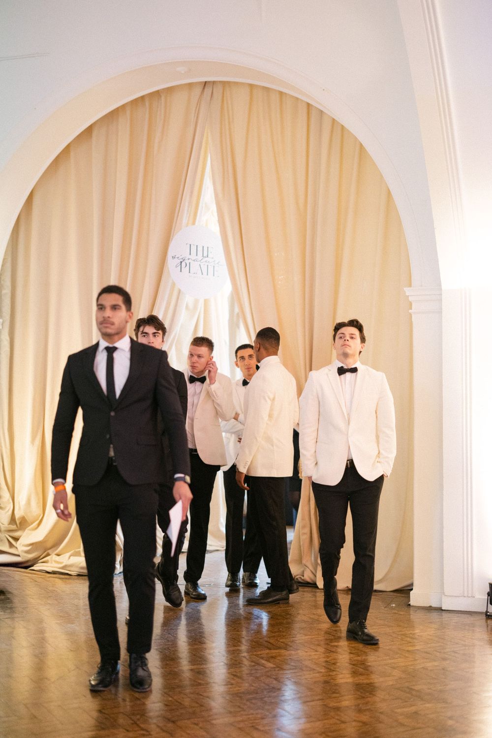 Groomsmen in black and white tuxedos walking through a draped archway at an elegant wedding reception venue.