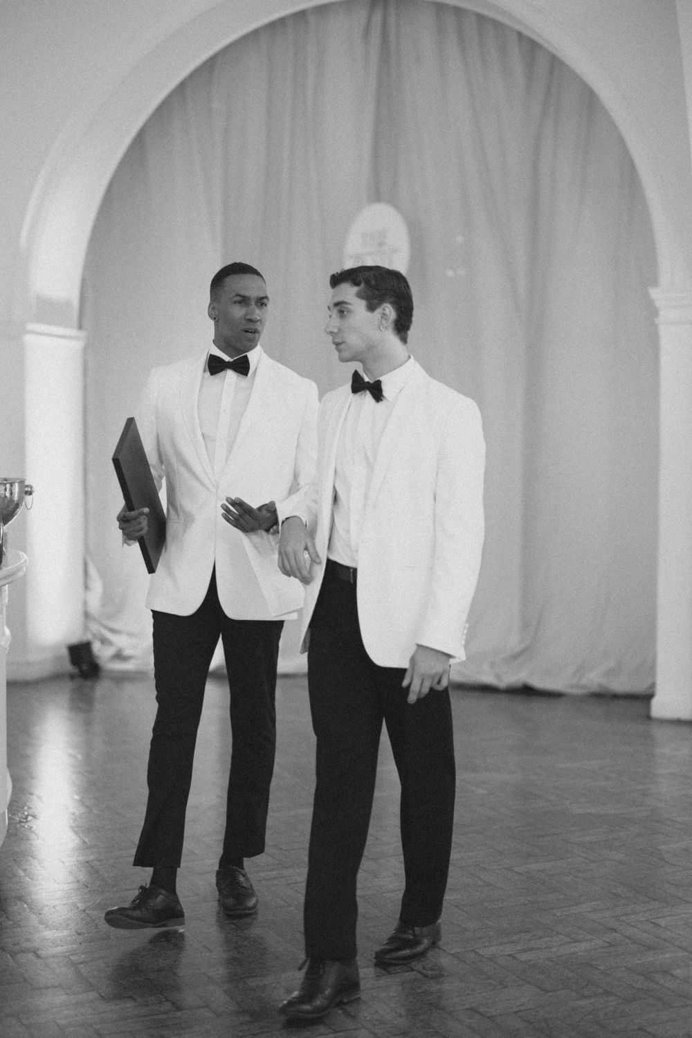 Two men in white tuxedo jackets and black bow ties conversing in an elegant ballroom, black and white photo.