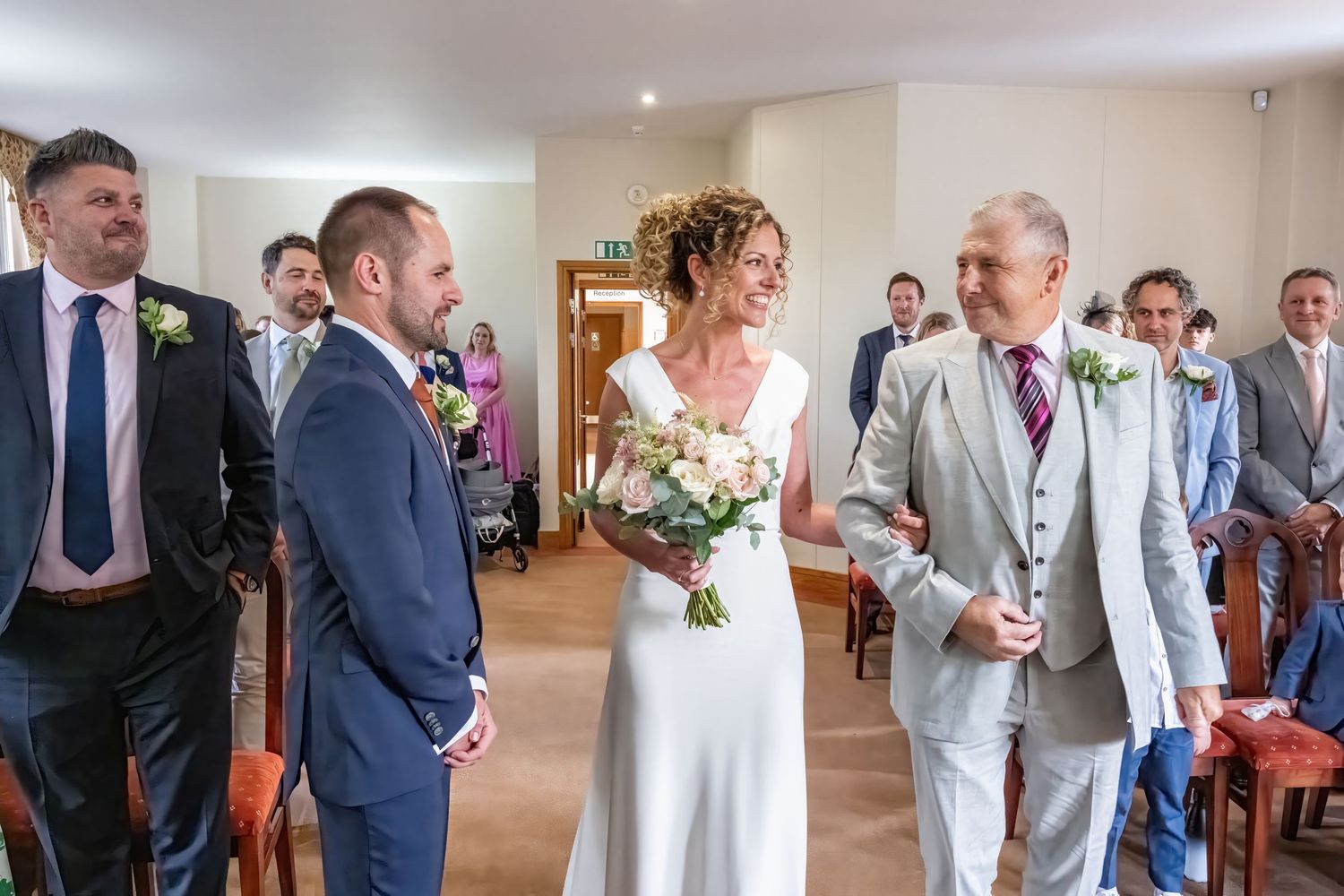 Bride walks down the aisle with older gentleman at an indoor wedding ceremony, greeted by smiling groom in navy suit.