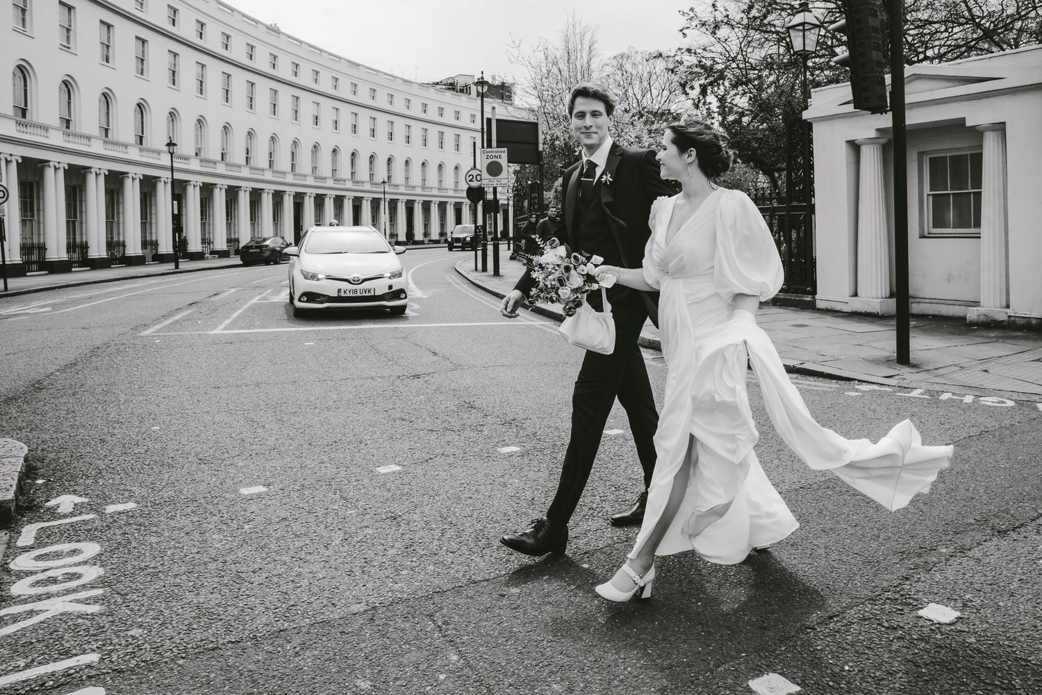 Bride and groom crossing a street in black and white wedding photo with elegant Georgian buildings in background.