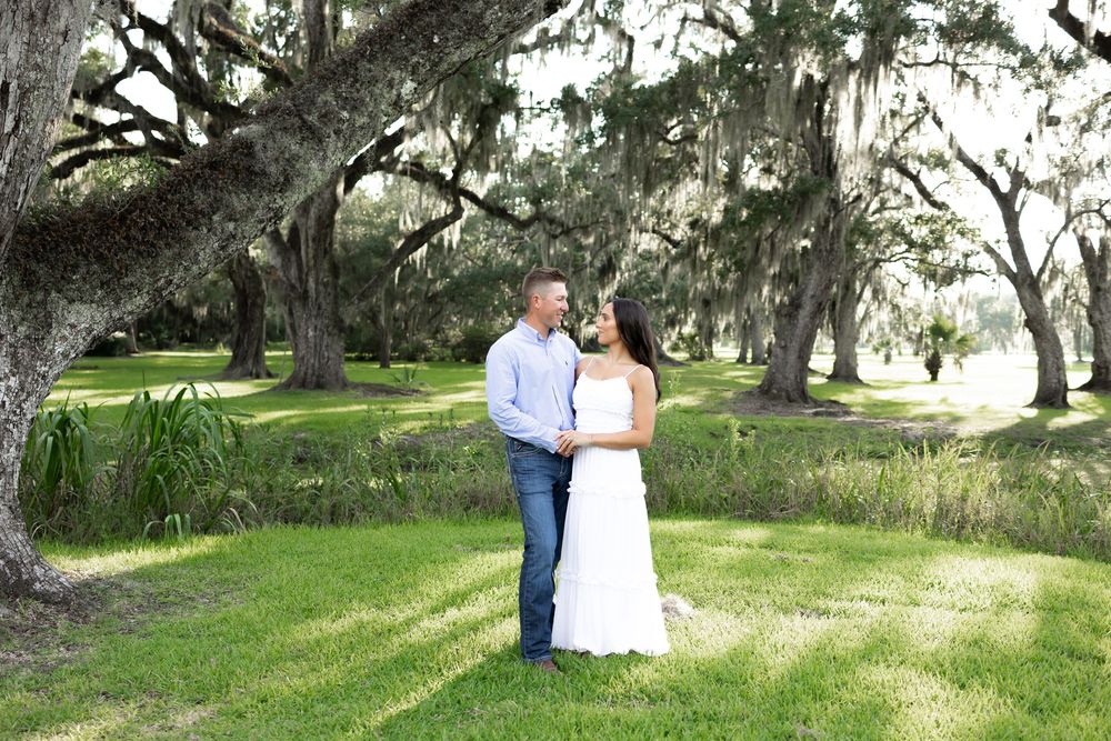 Couple sharing a romantic moment under sprawling oak trees draped with Spanish moss on a lush green lawn.