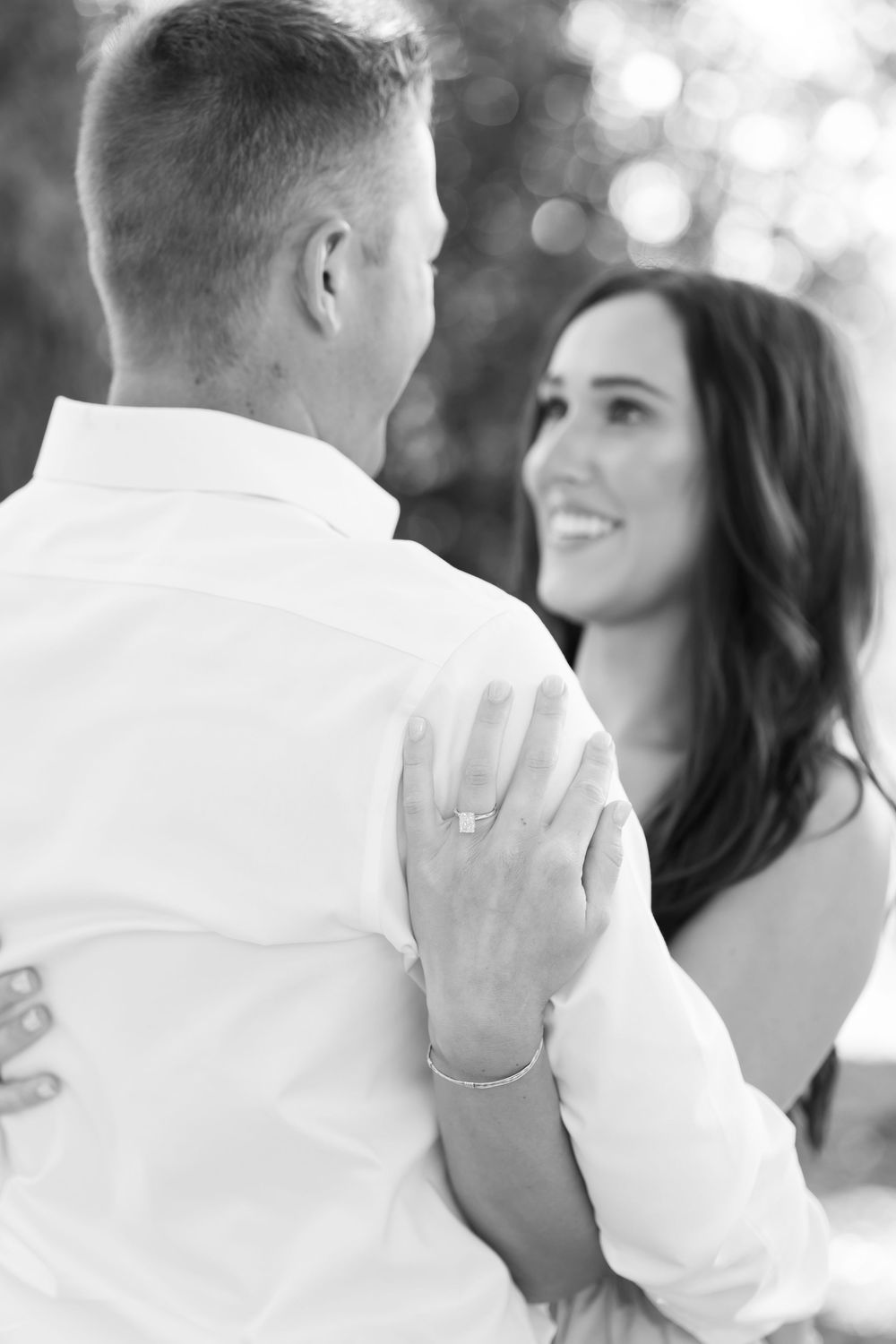 Black and white photo of smiling bride showing engagement ring while embracing groom outdoors.