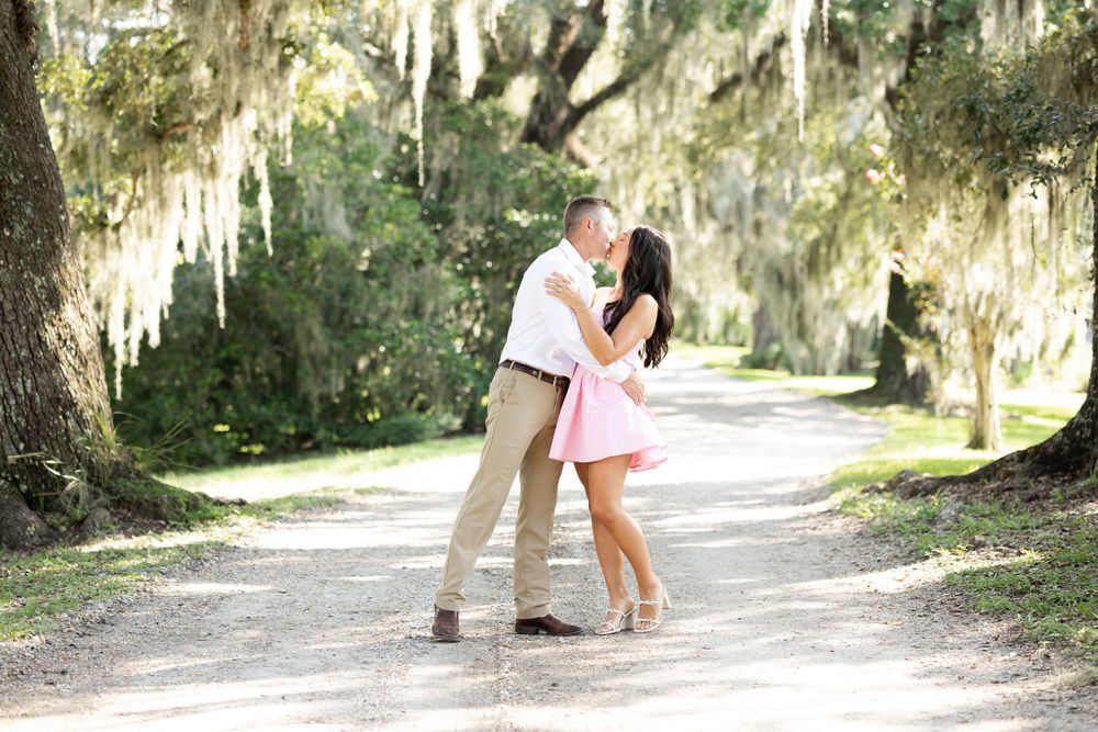Couple sharing a romantic kiss on a tree-lined path draped with Spanish moss in a lush outdoor setting.