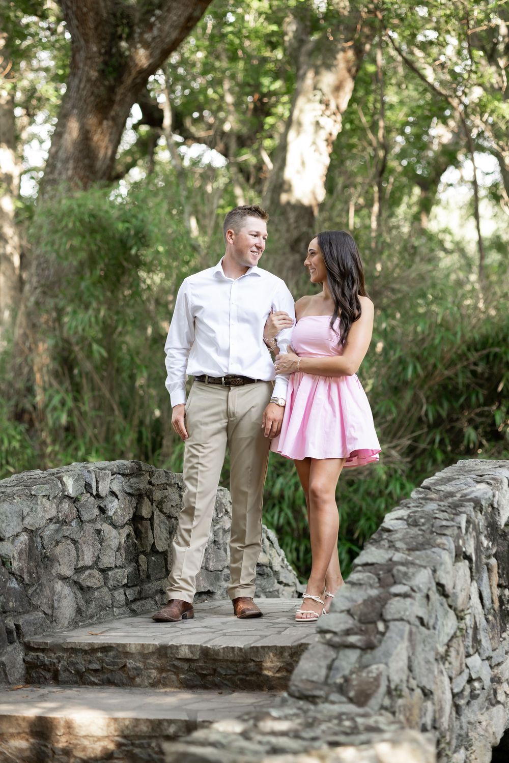 Happy couple walking hand-in-hand on a stone bridge surrounded by lush green trees.
