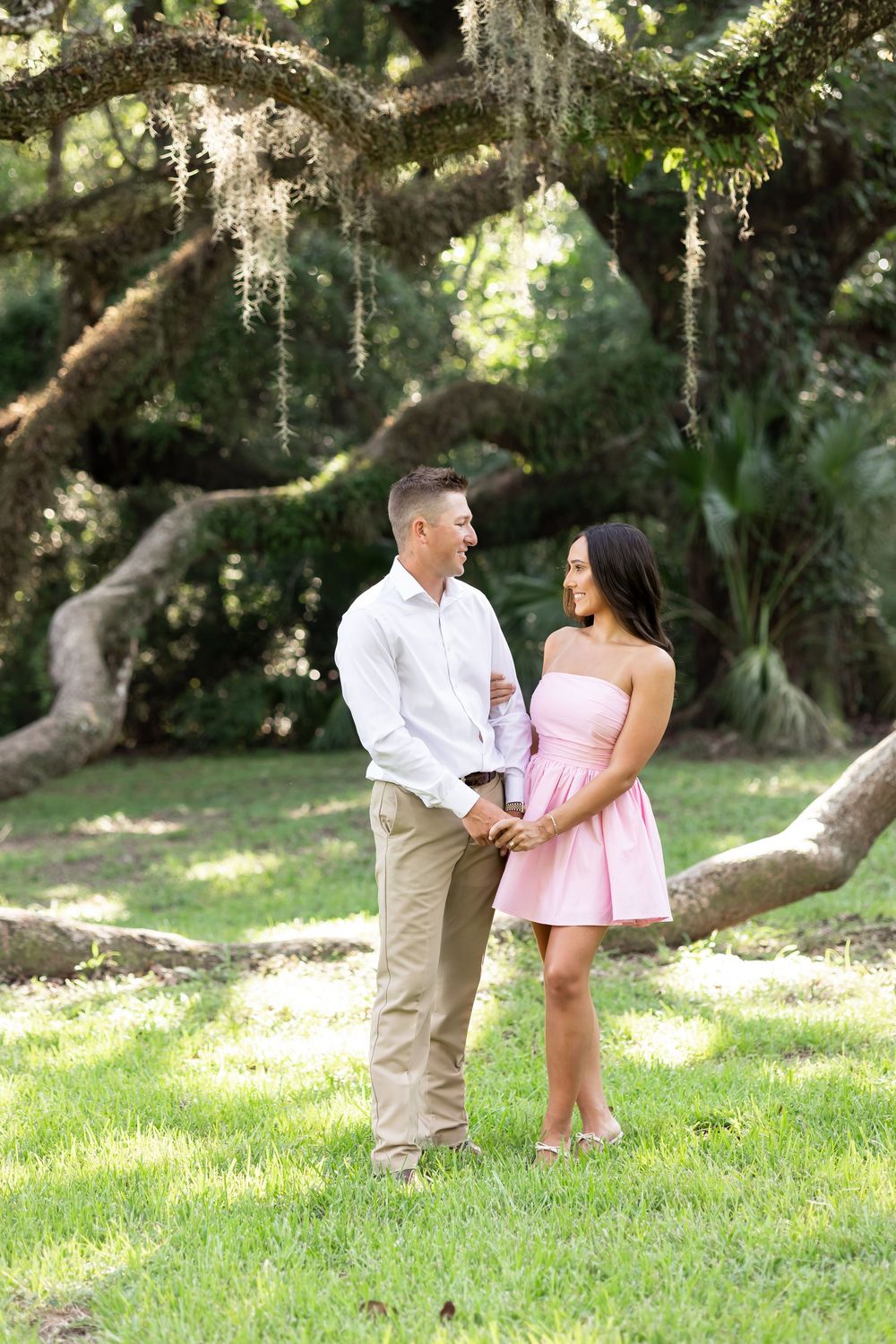 Couple holding hands and smiling at each other in a lush green park under a large oak tree with Spanish moss.