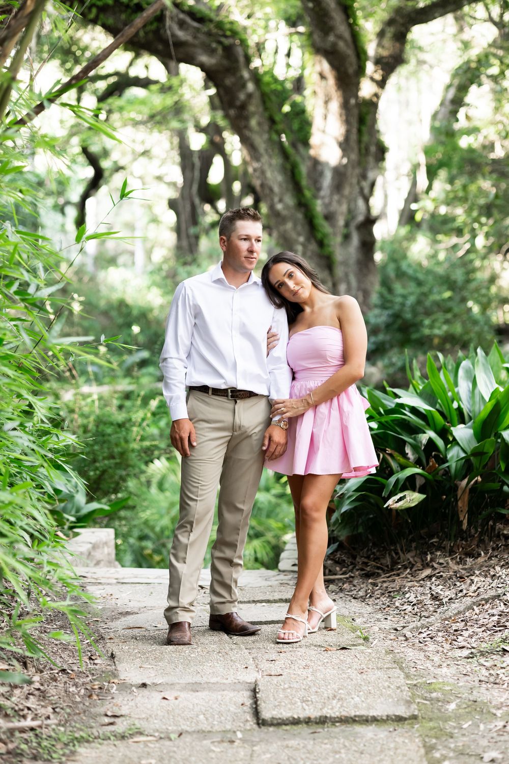 Couple walking hand in hand on a garden path surrounded by lush greenery, woman in pink dress, man in white shirt.