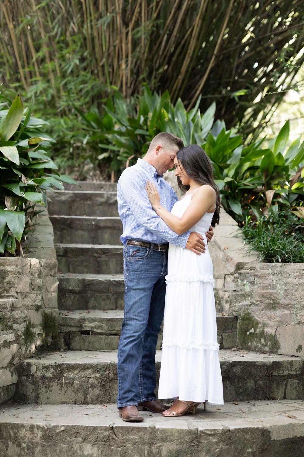 Romantic couple sharing an intimate moment on stone steps in a lush garden, woman in white dress, man in blue shirt.