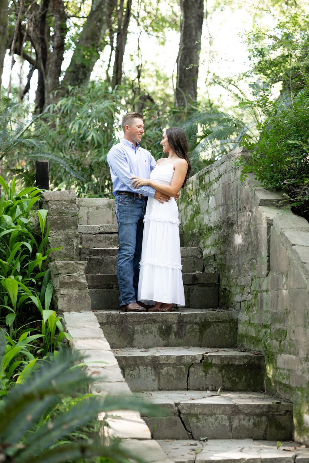 Couple embracing and smiling on stone steps surrounded by green foliage, woman in white dress, man in blue shirt.