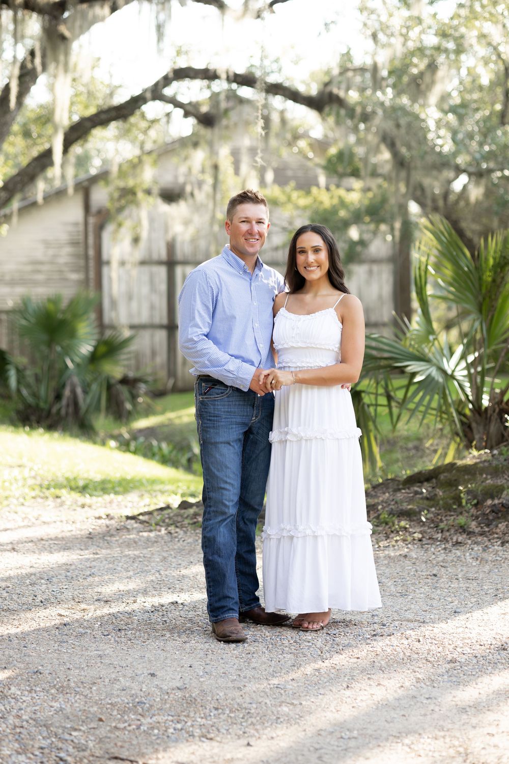 Couple posing outdoors on gravel path under Spanish moss oak tree, man in blue shirt and jeans, woman in white maxi dress.