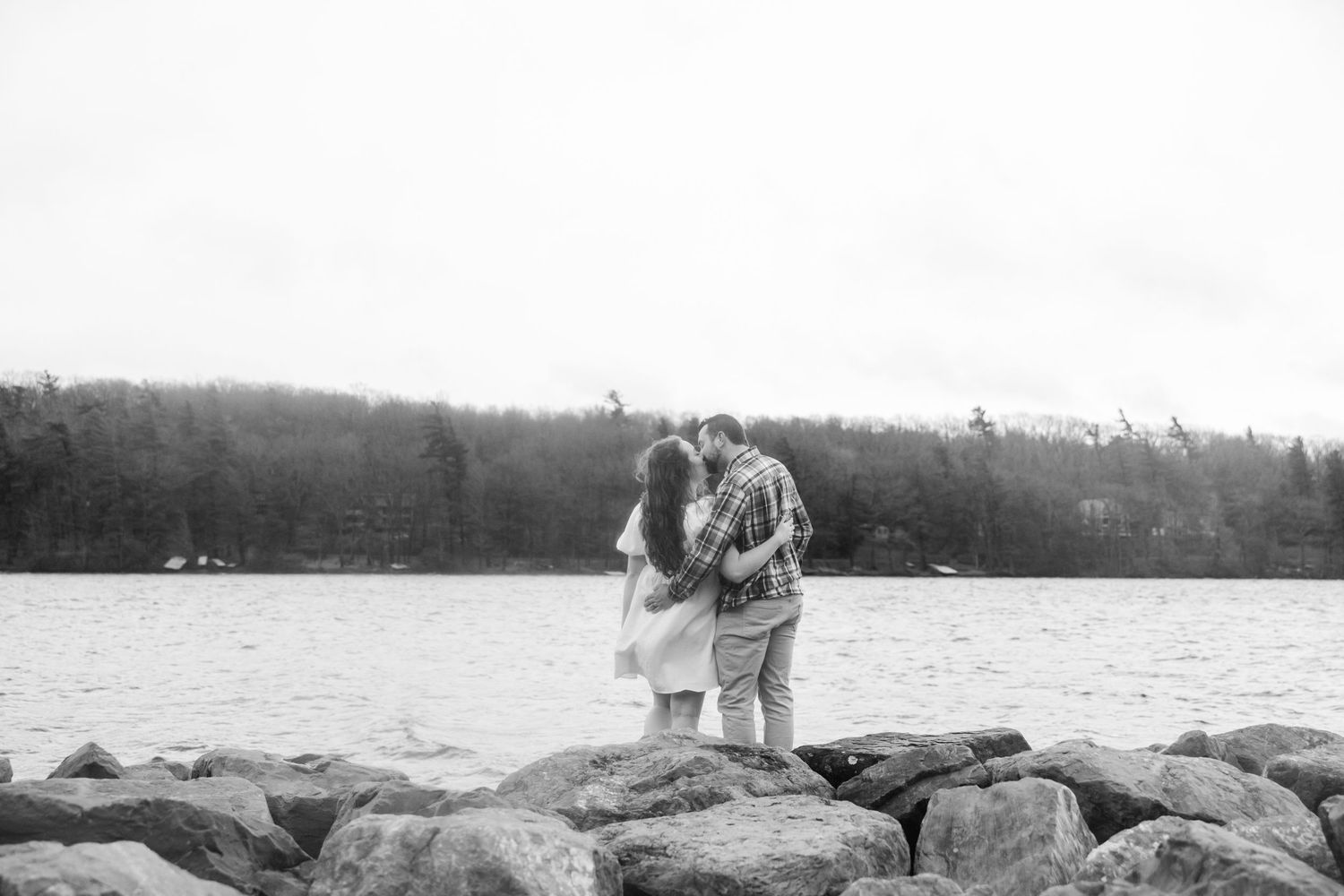 Couple embracing on rocky shoreline in black and white, with forested hills and calm water in background. deep creek lake state park md