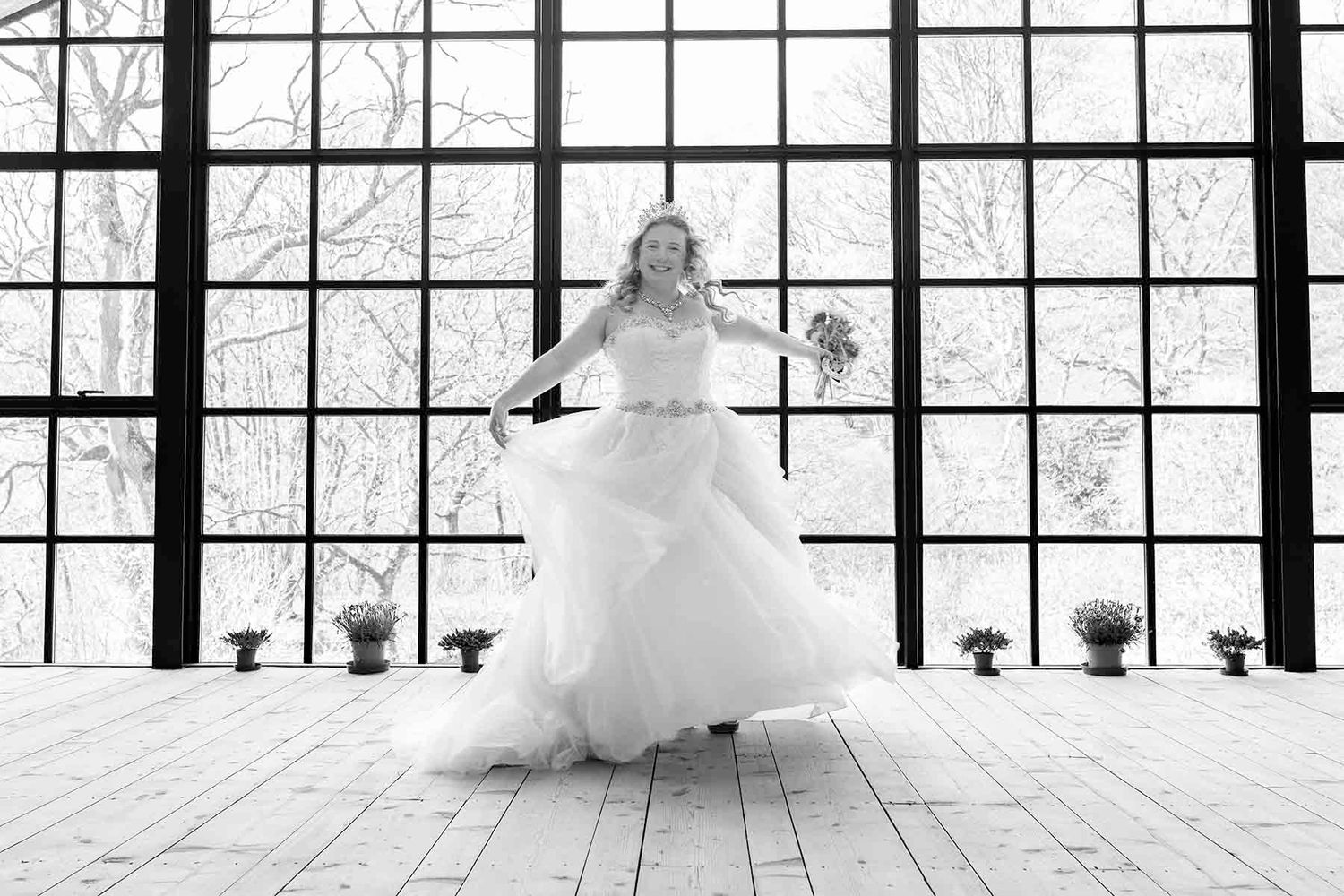 Bride in white ball gown twirling in sunlit industrial venue with large windows, black and white wedding photo.