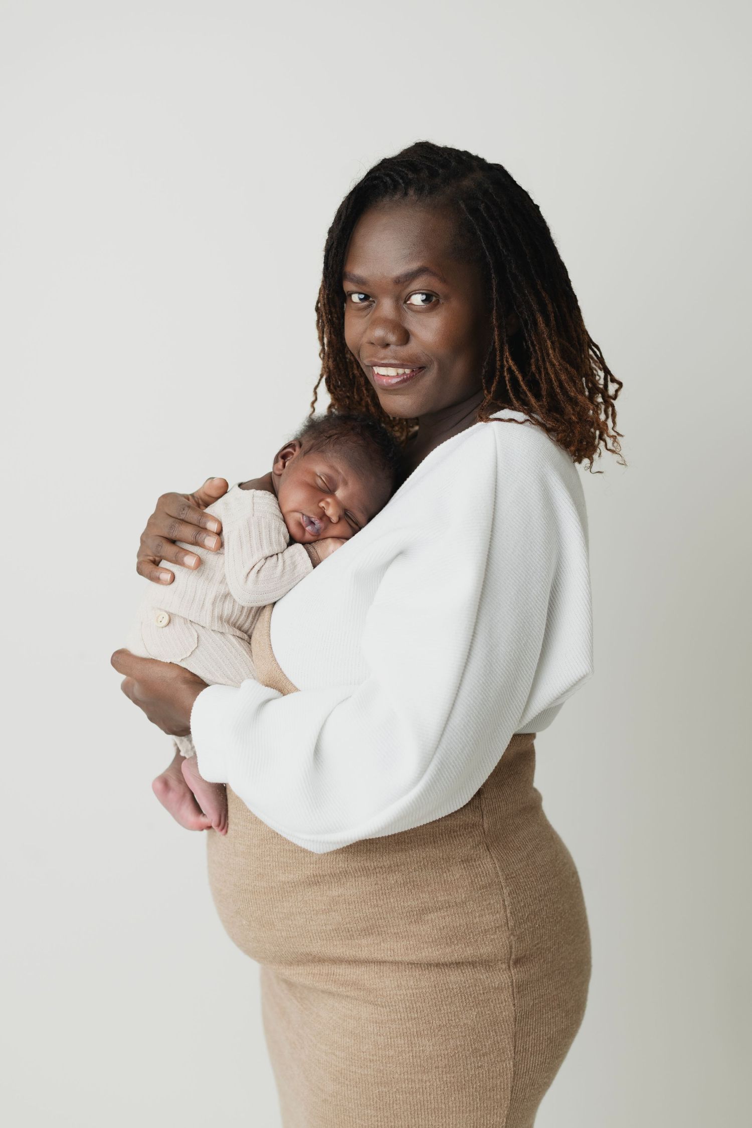 A smiling Black mother holds her sleeping newborn baby against her chest in a bright studio portrait. Elizabeth Lang Photography Surrey