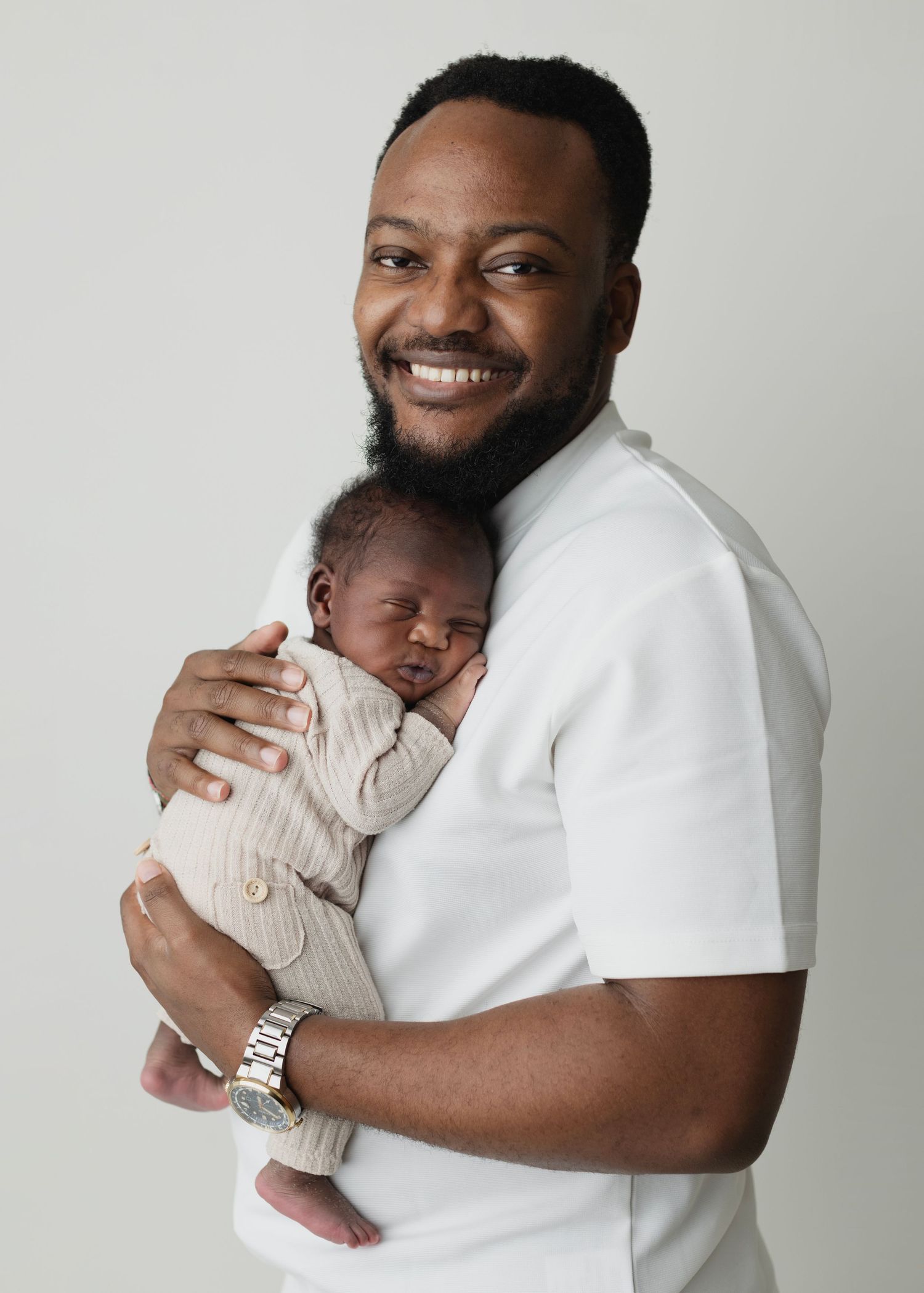 Smiling man in white shirt holding a sleeping newborn baby, studio portrait on light gray background. Elizabeth Lang Photography Surrey