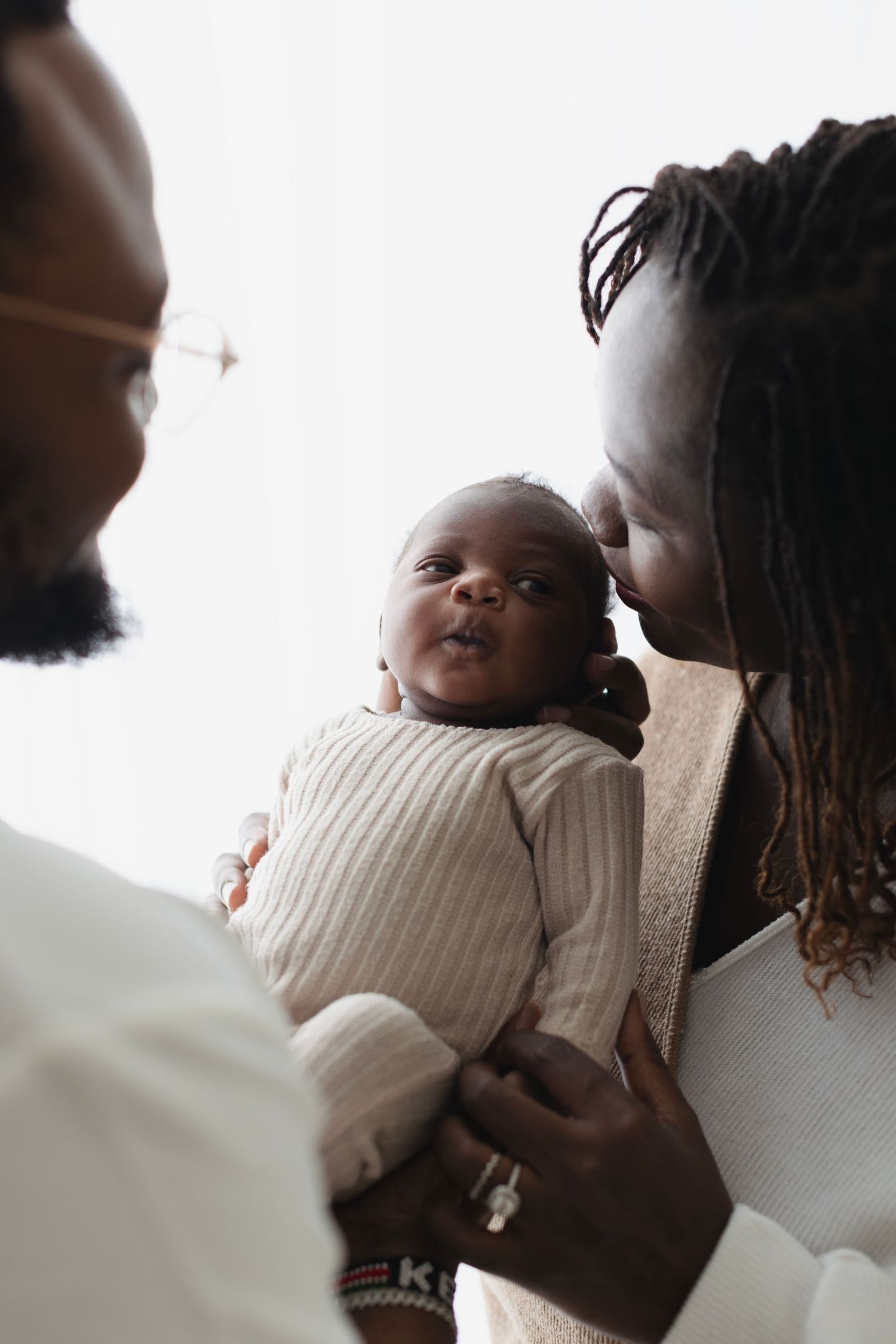 Black parents share a tender moment with their alert newborn baby in a softly lit portrait. Elizabeth Lang Photography Surrey