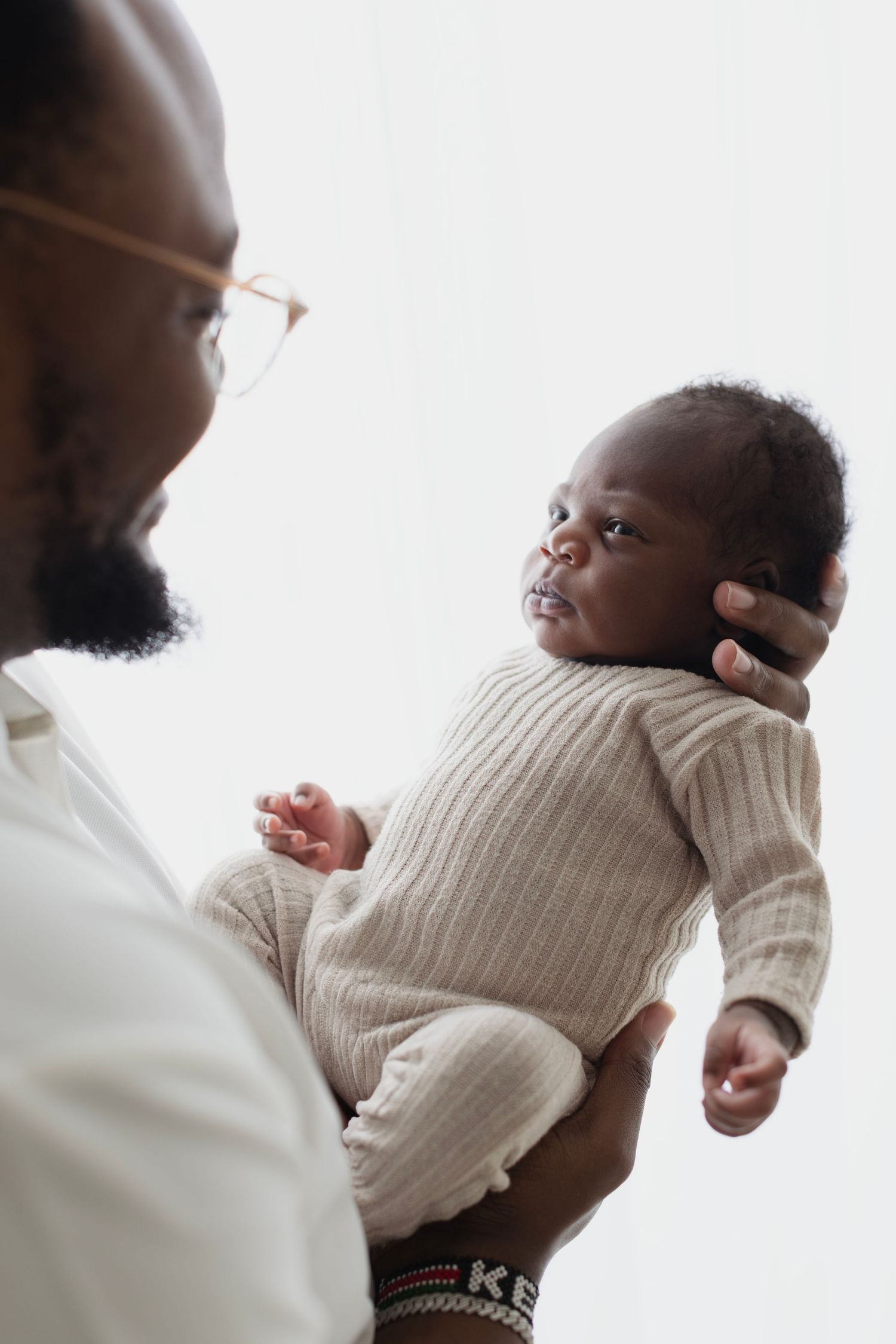 A Black father tenderly holds his newborn baby dressed in a beige knit onesie, sharing a gentle moment together. Elizabeth Lang Photography