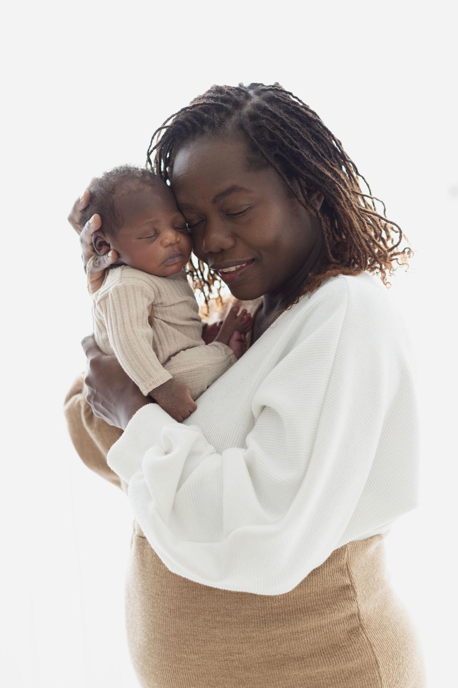 A smiling Black mother tenderly holds her newborn baby close to her cheek against a white background. Elizabeth Lang Photography Surrey