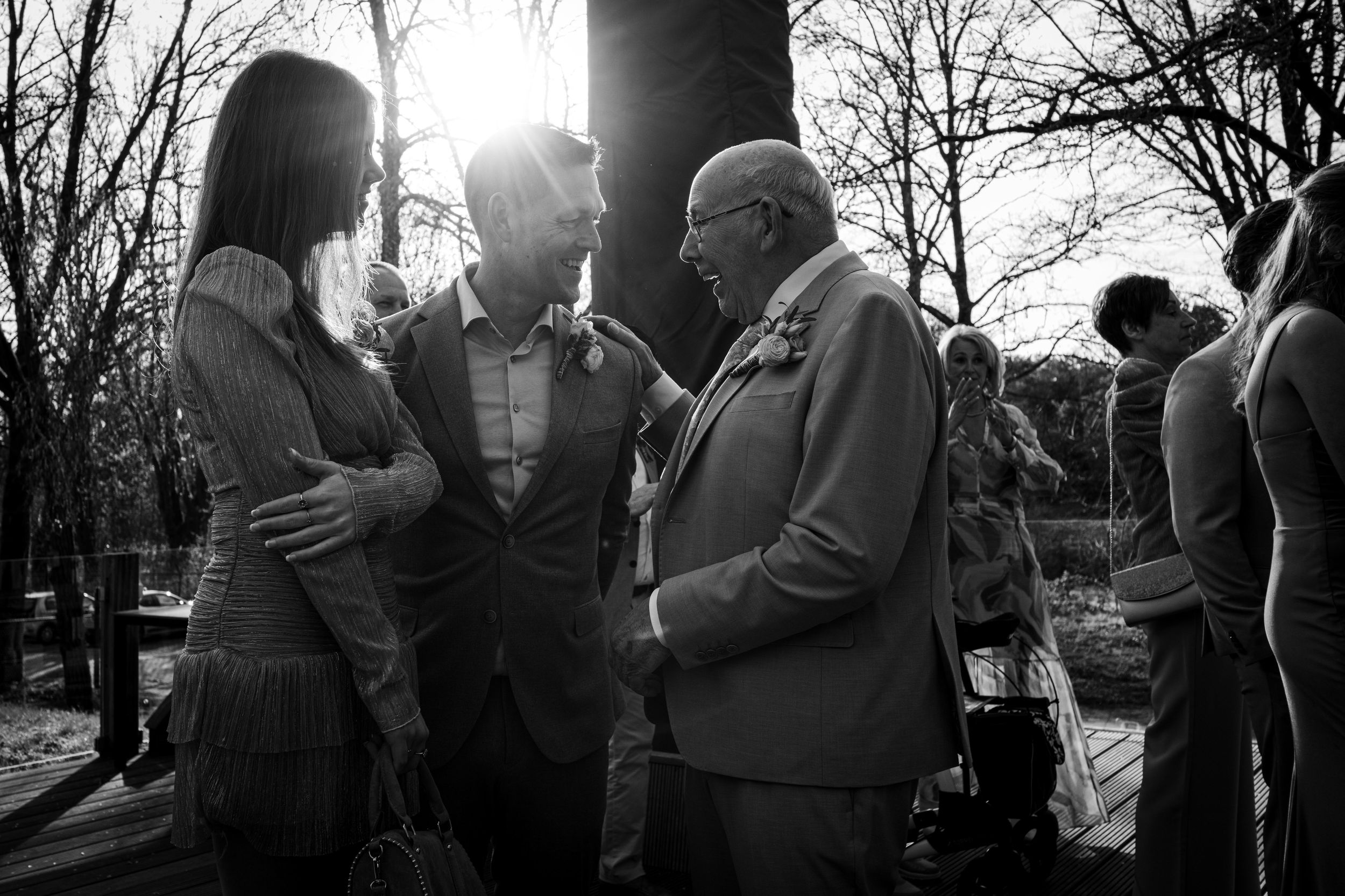 Black and white photo of guests conversing at an outdoor wedding reception with sunlight filtering through bare trees.