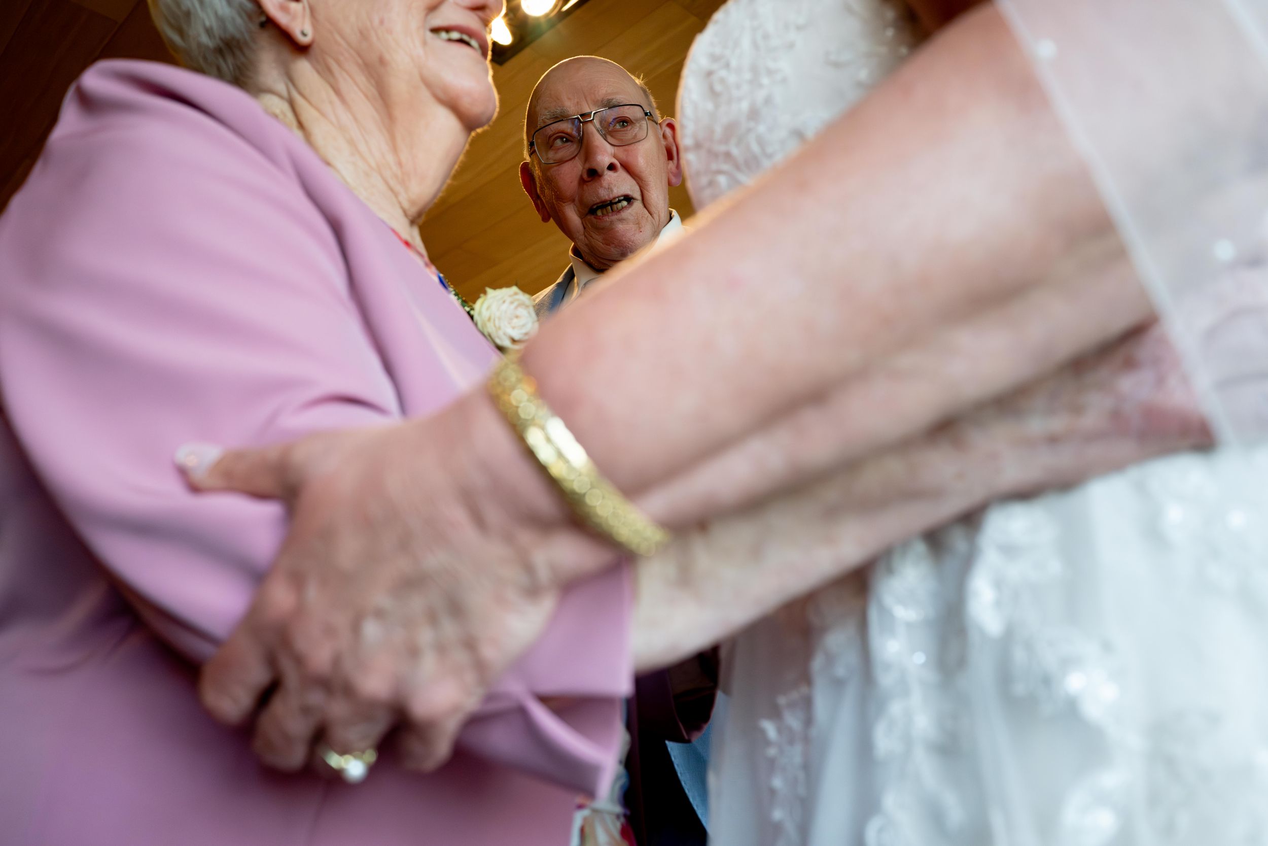 Elderly couple joyfully greeting a bride in a white wedding gown at a wedding reception celebration.