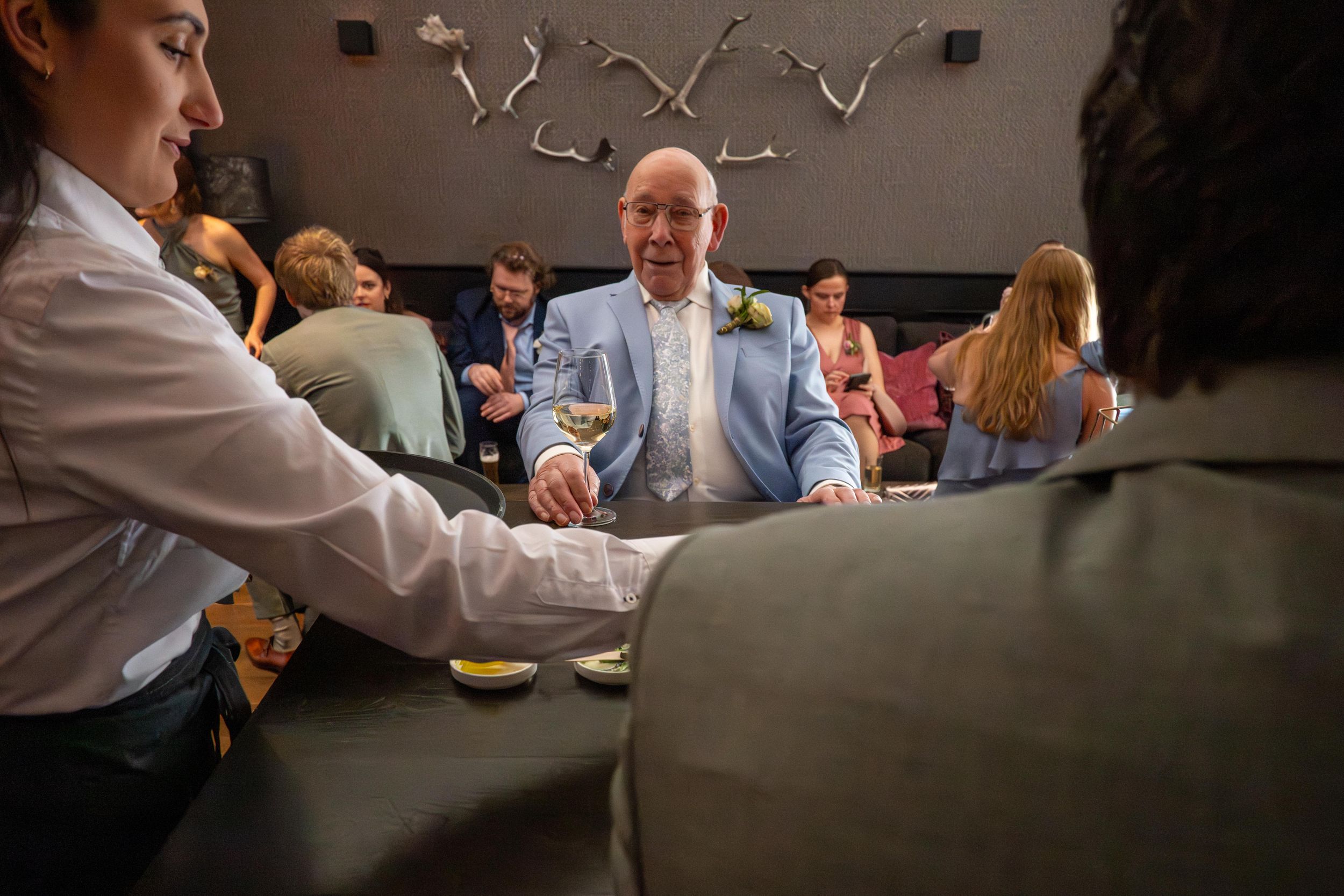 Elegantly dressed man in light blue suit holding wine glass at a busy wedding reception venue.