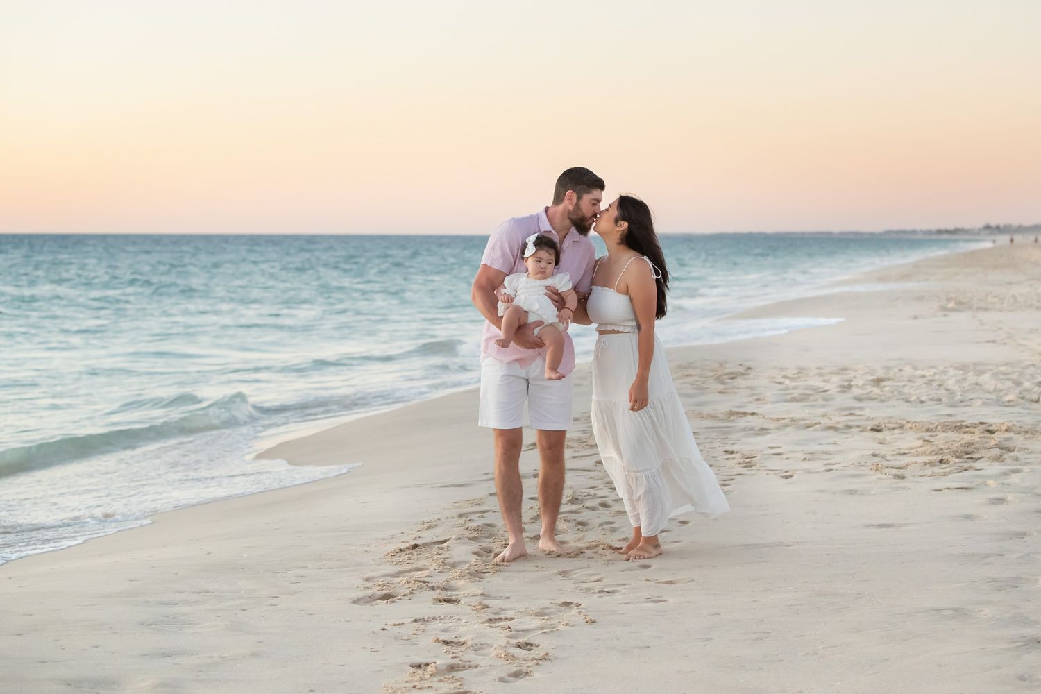 Happy family walking on beach at sunset, father holding baby while parents share a kiss.