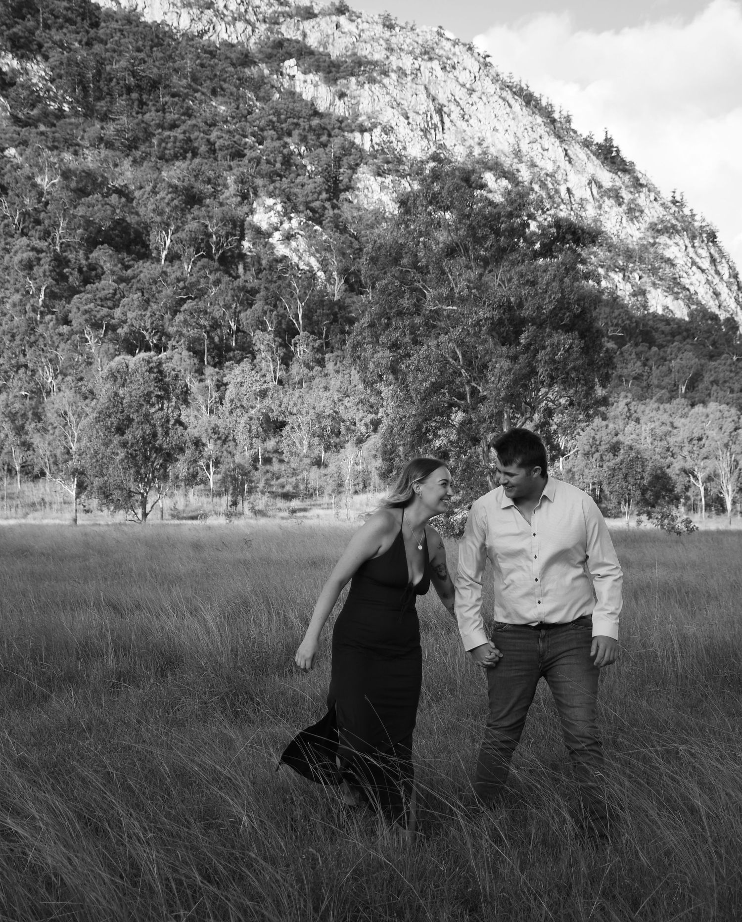 Black and white photo of a couple walking together in a grassy field with mountain backdrop.
