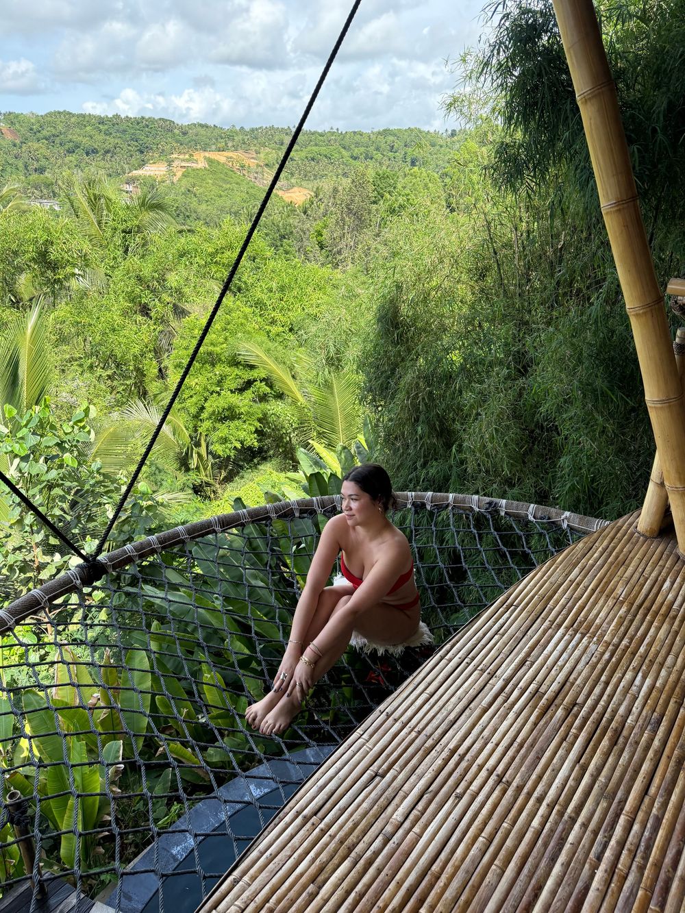 Woman in bikini relaxing on a bamboo net perch overlooking a lush tropical jungle valley in Bali, Indonesia.