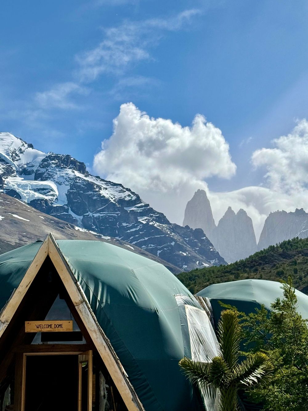 Green glamping dome with wooden A-frame entrance and dramatic snow-capped Torres del Paine peaks behind.