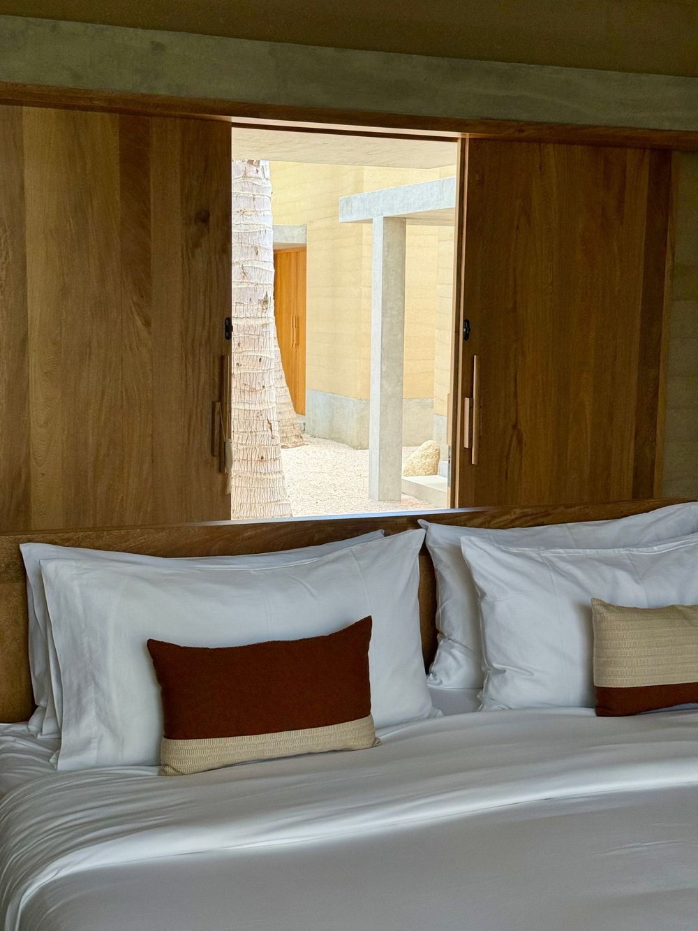 Elegant hotel bedroom with white bedding, terracotta accent pillows, wooden headboard, and a window overlooking a sunlit courtyard.