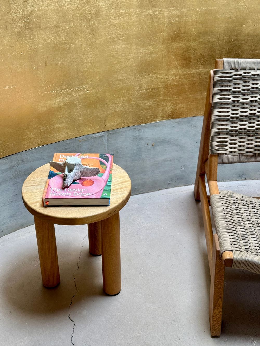 Minimalist wooden side table with books and a woven lounge chair on a concrete floor against a golden wall.
