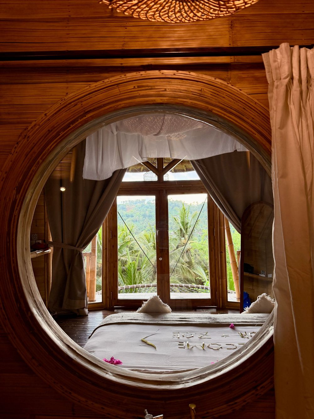 Circular wooden window frame revealing a cozy bed with welcome flower petals in a Balinese jungle villa.