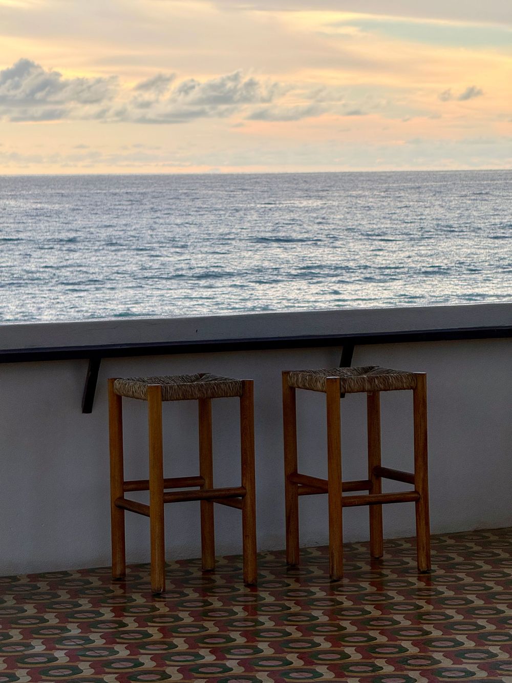 Two wooden bar stools on a patterned terrace overlooking a calm ocean at sunset with colorful clouds.