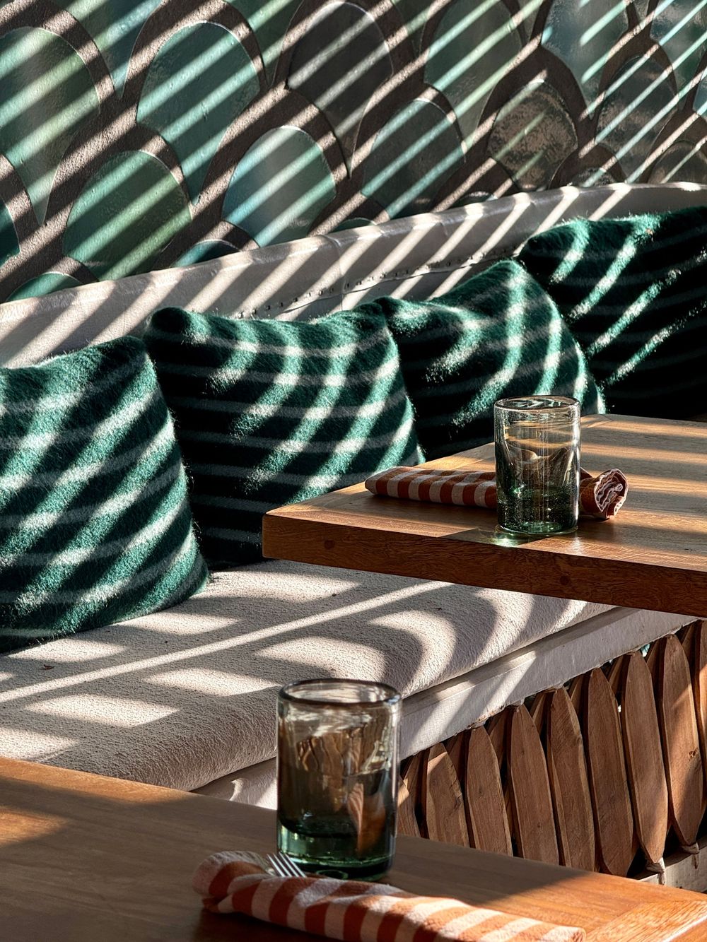 Outdoor lounge with green striped cushions casting dramatic shadows, wooden table with green glasses in sunlight.