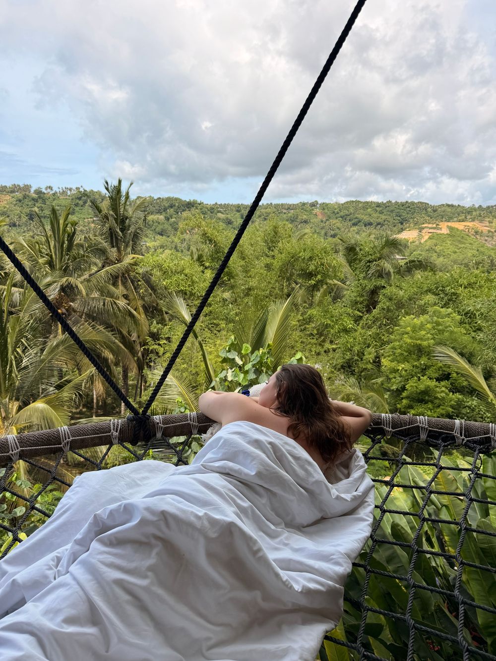 Woman relaxing in hanging net bed wrapped in white sheets overlooking lush tropical jungle canopy in Bali.