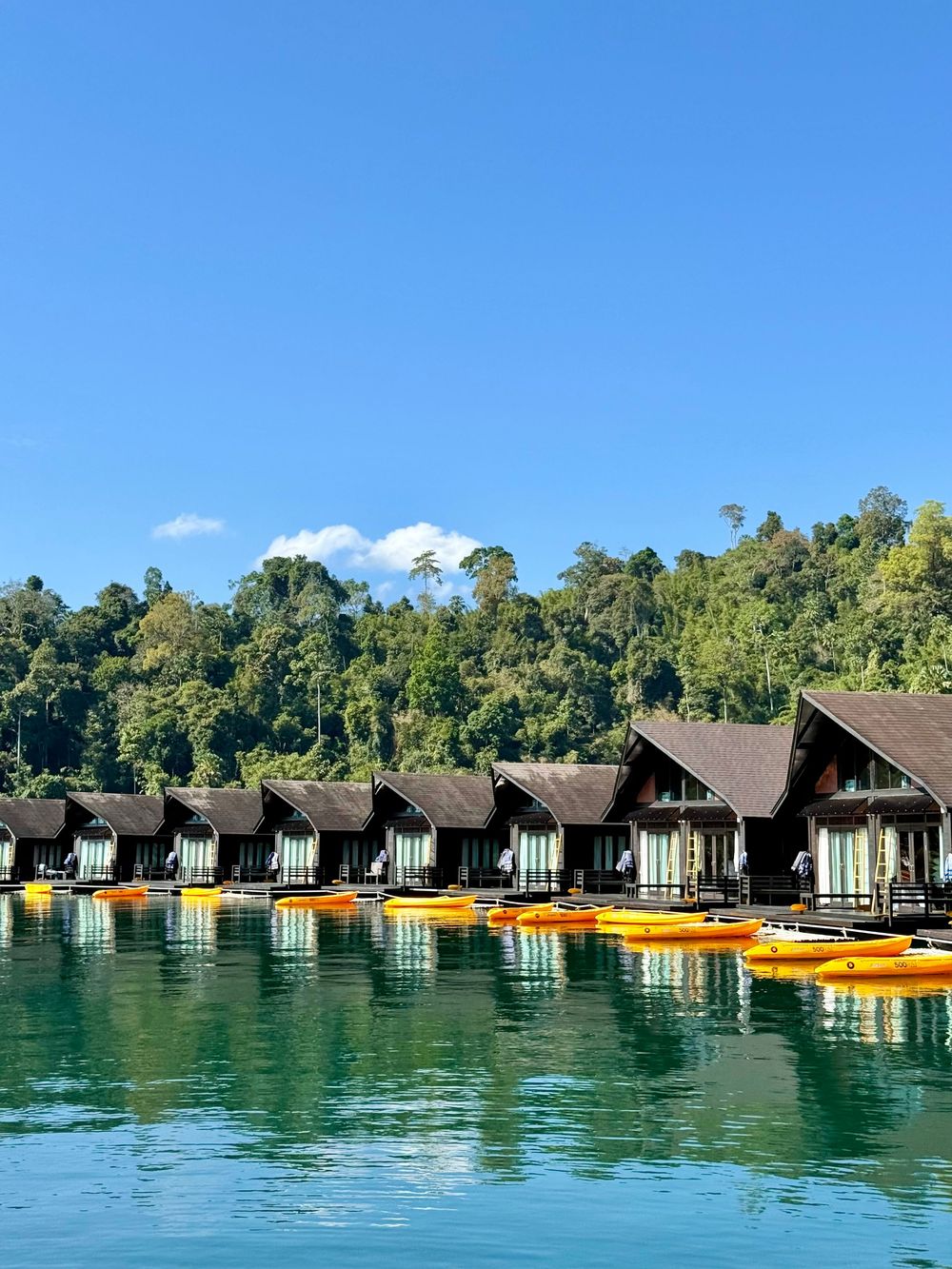 Overwater bungalows with orange kayaks reflecting in turquoise lake with forested mountains backdrop in Khao Sok National Park, Thailand.