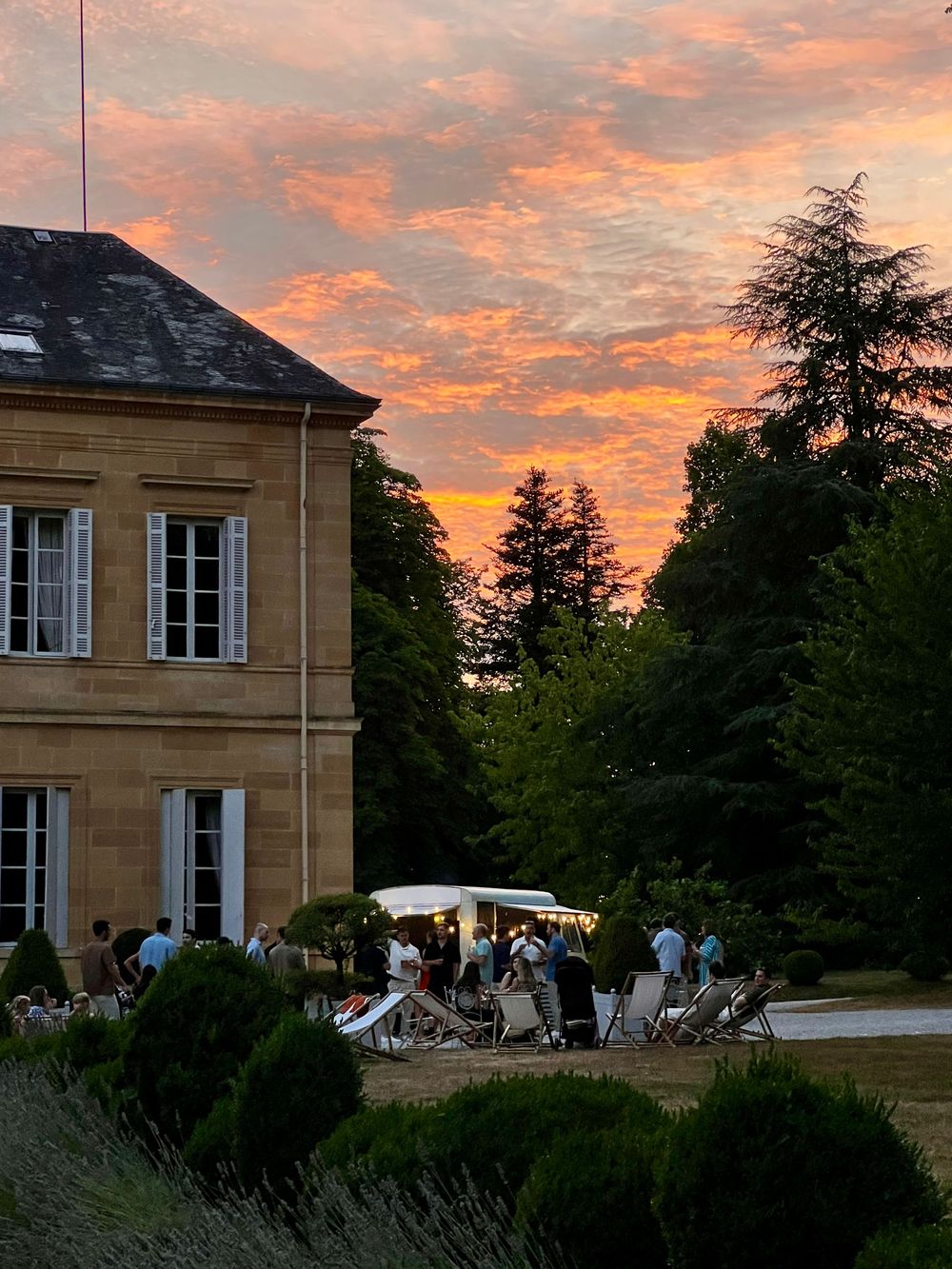 Golden sunset sky with vibrant orange and pink clouds over an elegant French chateau garden with outdoor gathering.