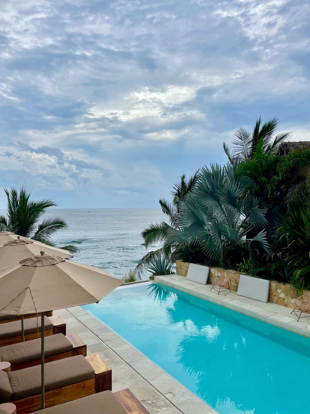 Infinity pool overlooking a turquoise ocean with palm trees and lounge chairs under cloudy skies.