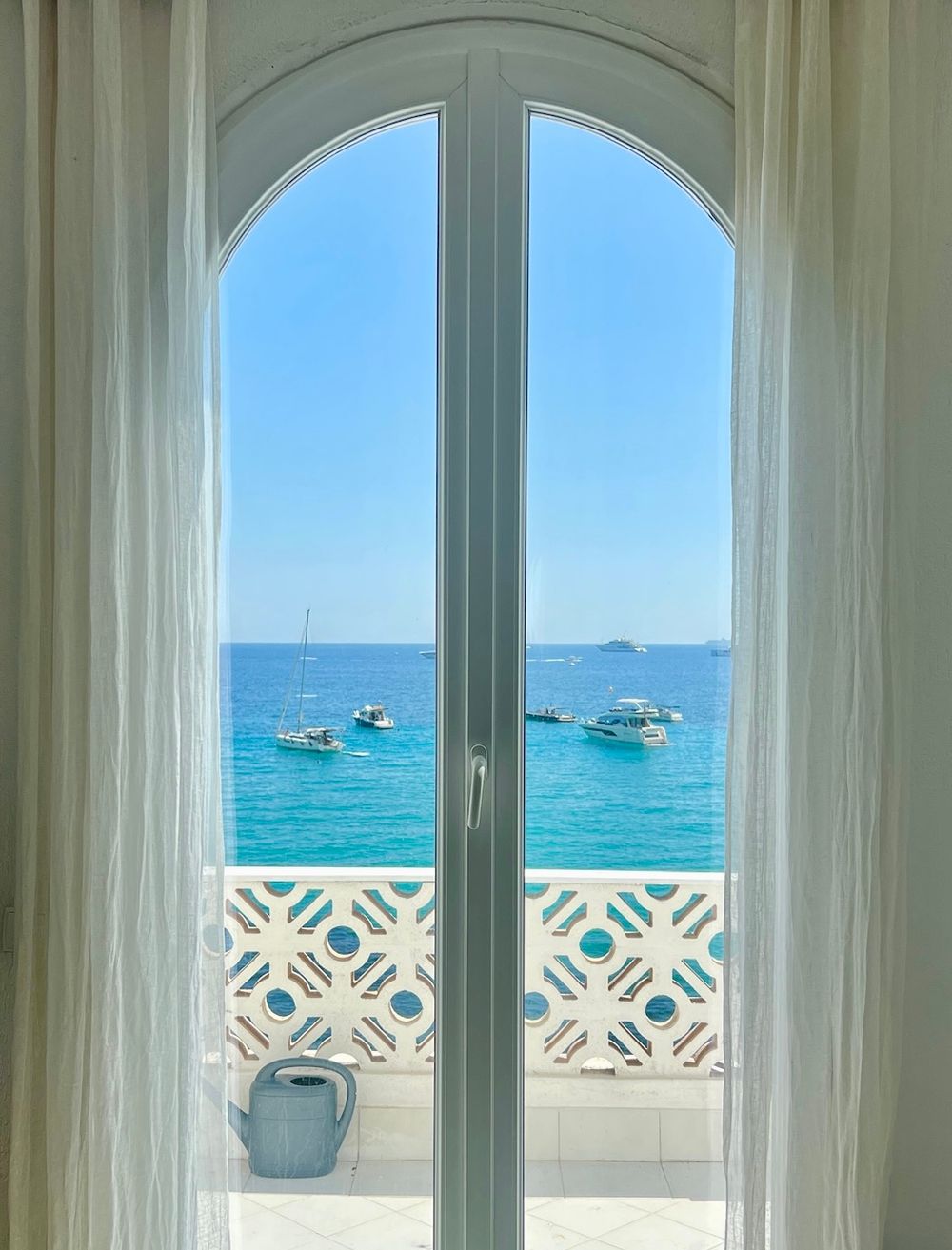 Arched French doors with white curtains framing a stunning turquoise ocean view with boats.