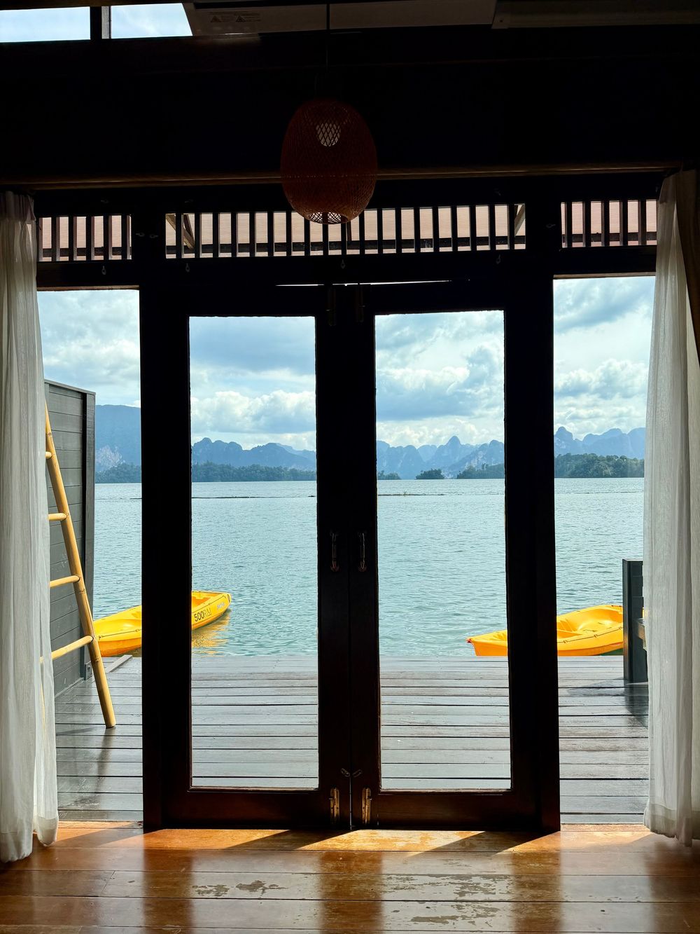View through wooden glass doors of a serene lake with karst mountains and yellow kayaks on a floating resort deck in Thailand.