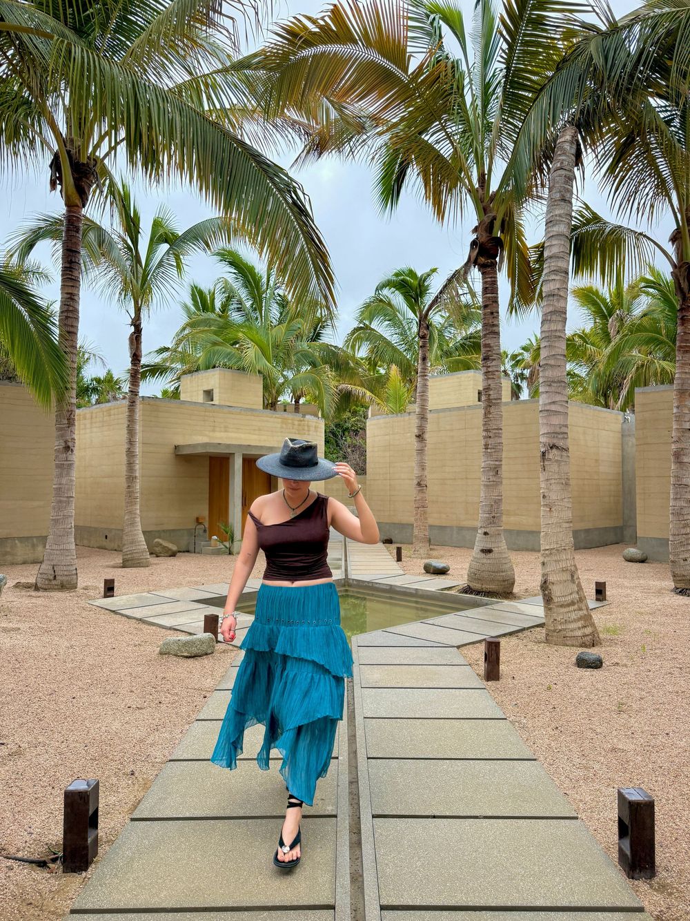 Woman in teal skirt and black hat walks along a tropical resort pathway lined with tall palm trees.