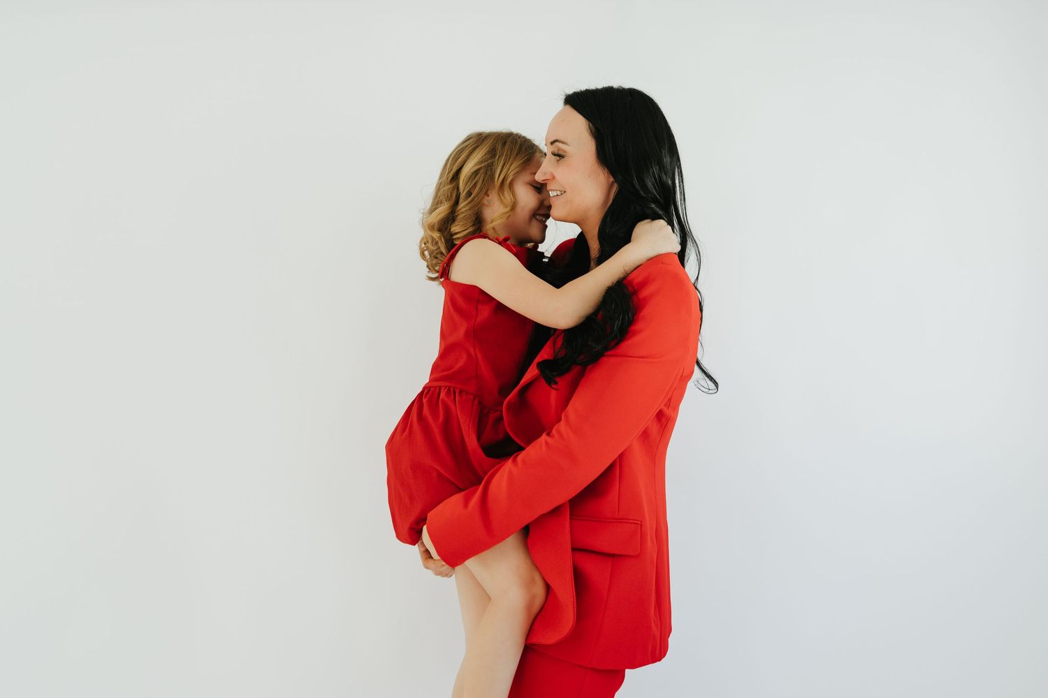 A woman in a red dress holds a young girl in red, sharing a joyful embrace against a white background.