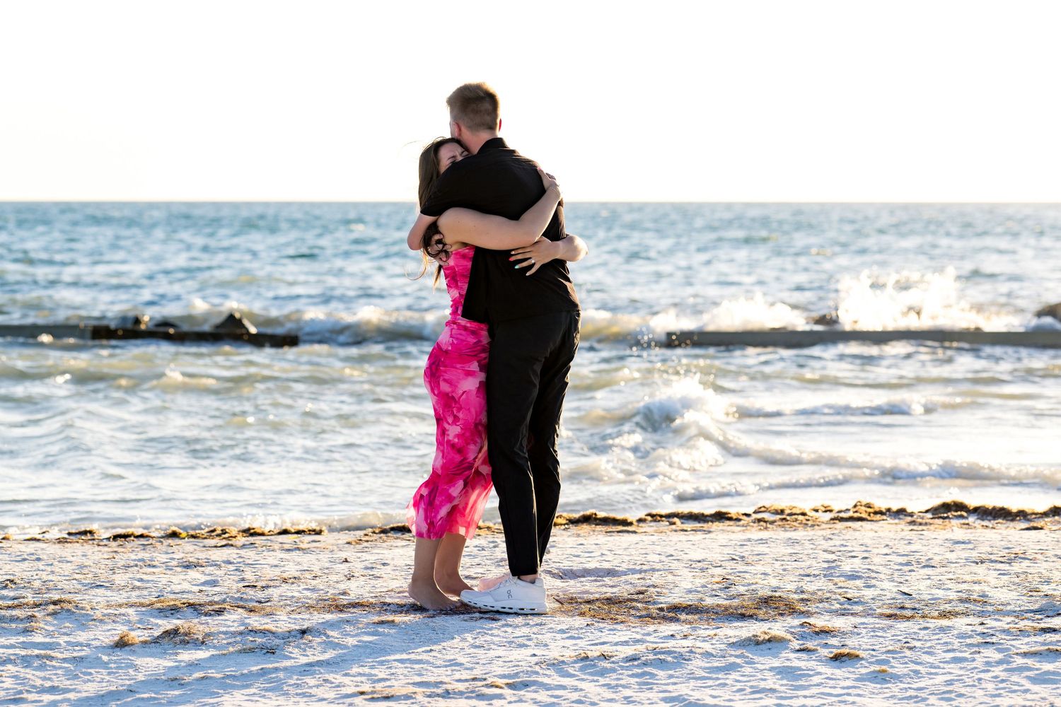 Couple embracing on a sandy beach at sunset with ocean waves in the background.