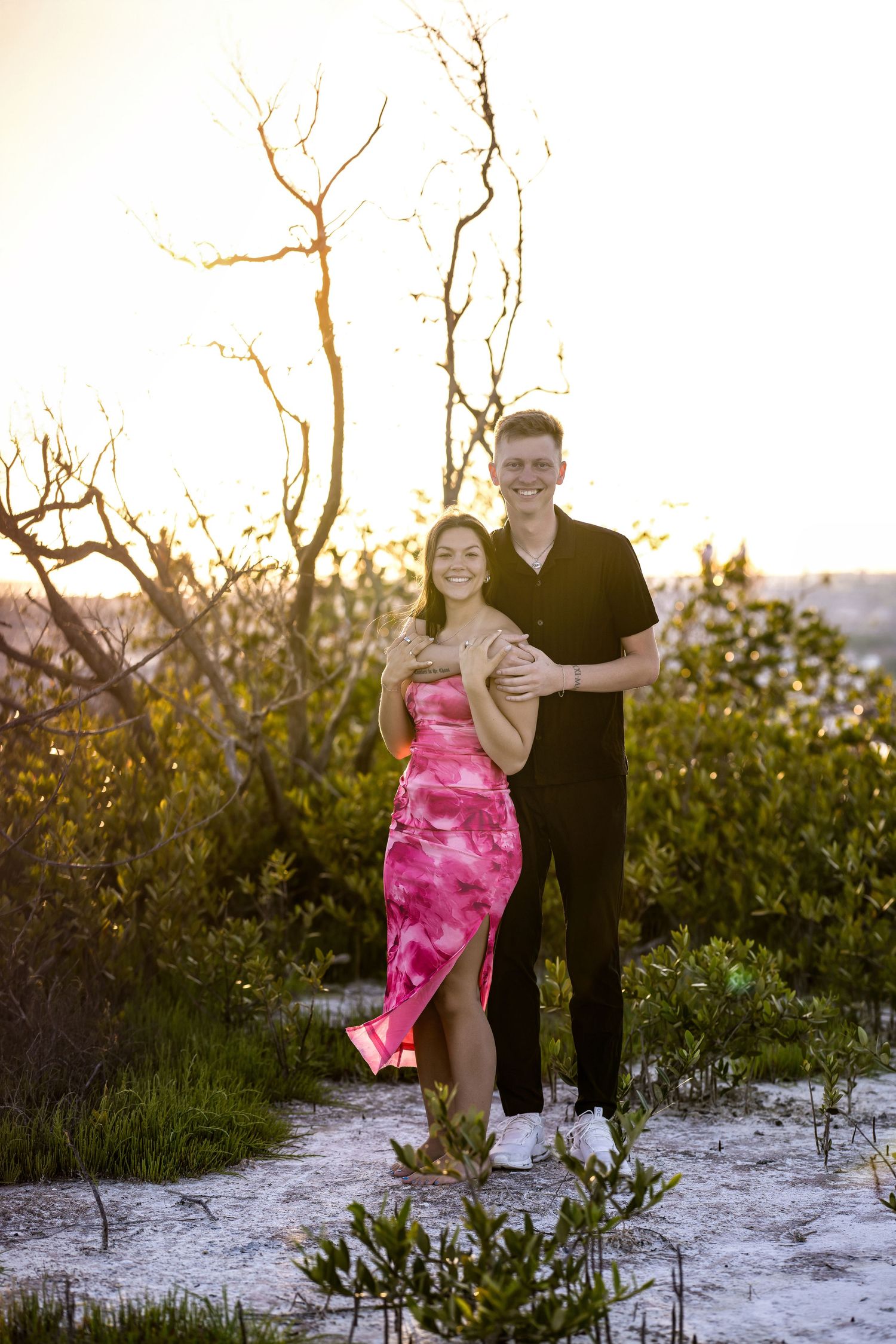 Couple posing outdoors at sunset, woman in pink tie-dye dress, man in black outfit, surrounded by lush greenery.