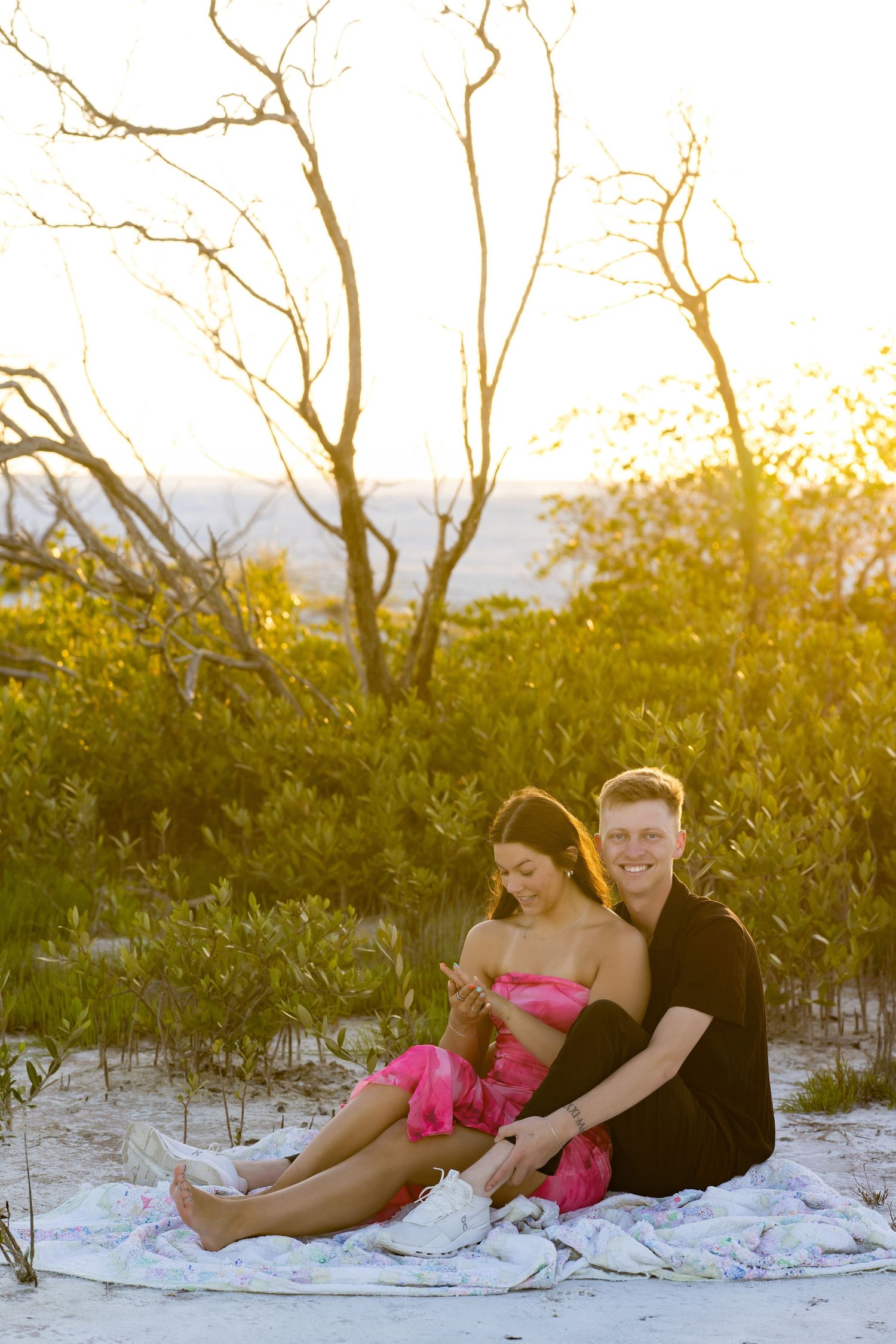 Couple sitting on a blanket at sunset on sandy beach, surrounded by lush greenery, woman in pink dress, man in black.