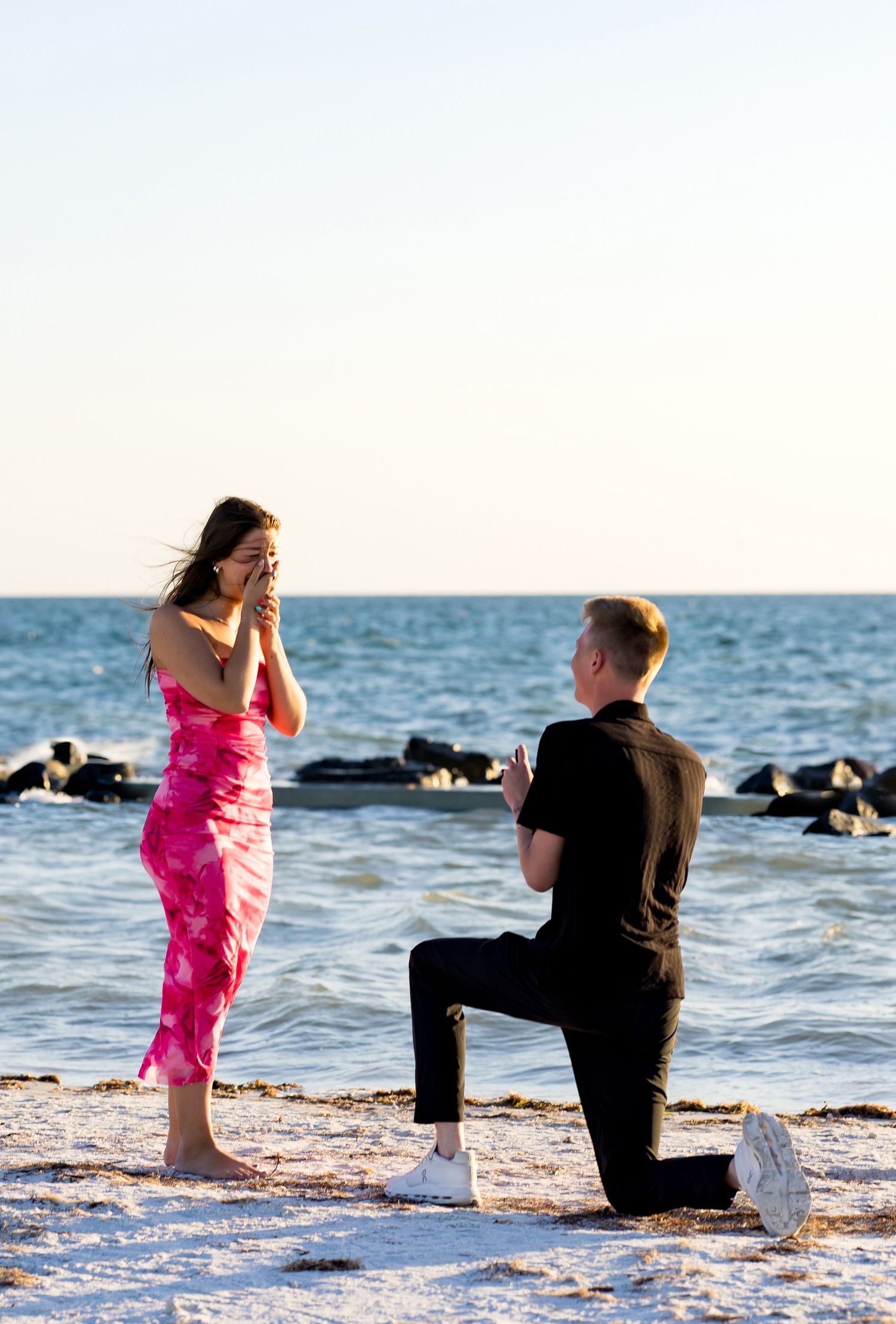 Man proposes on one knee to woman in pink dress on sandy beach at sunset by the ocean.