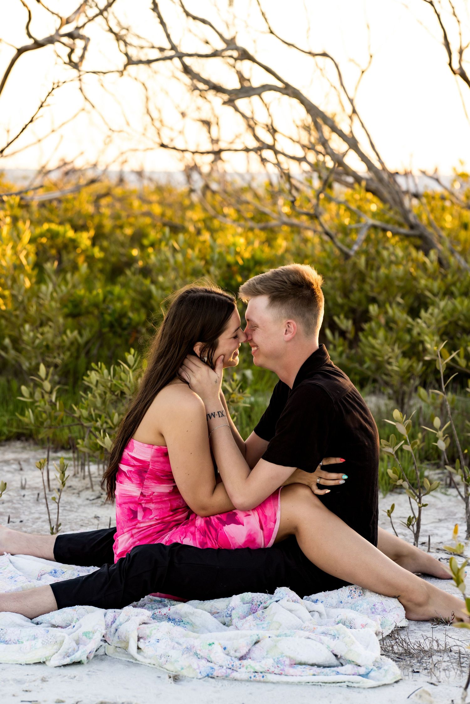Couple sitting on a blanket at the beach, leaning in for a kiss surrounded by lush greenery and golden hour light.