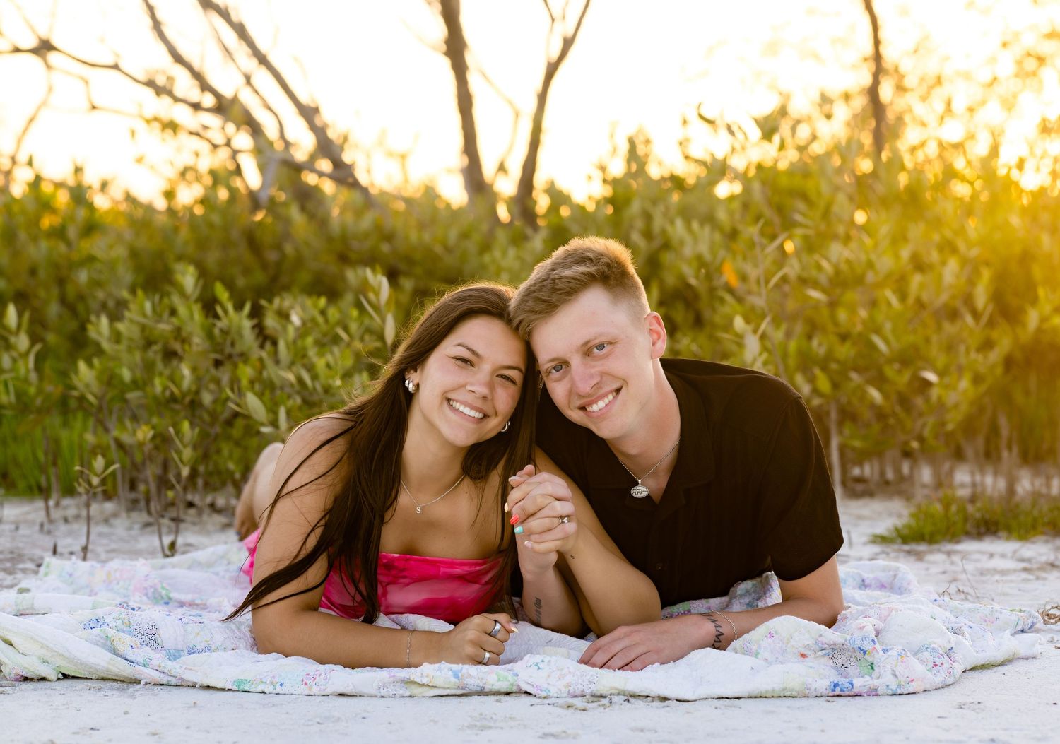 Happy couple lying on a blanket at a sandy beach during golden hour sunset with trees in background.